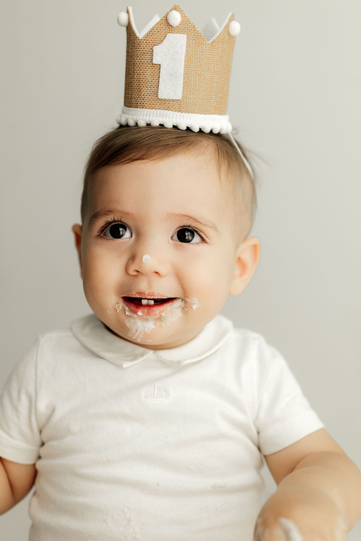 A baby wearing a white shirt and gold paper crown smiles with a messy face.