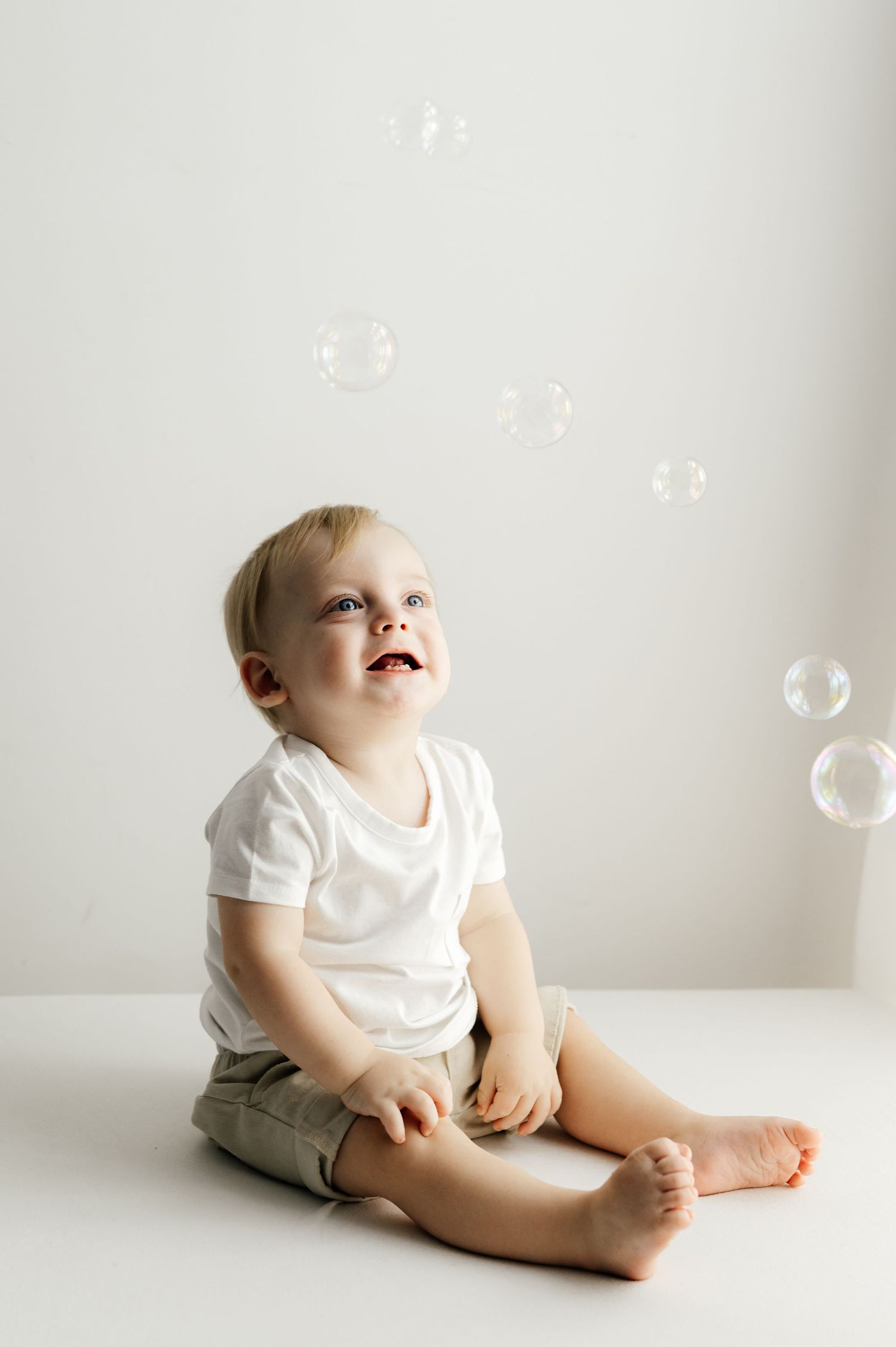 A toddler in white shirt and shorts sitting on floor watching soap bubbles float above.