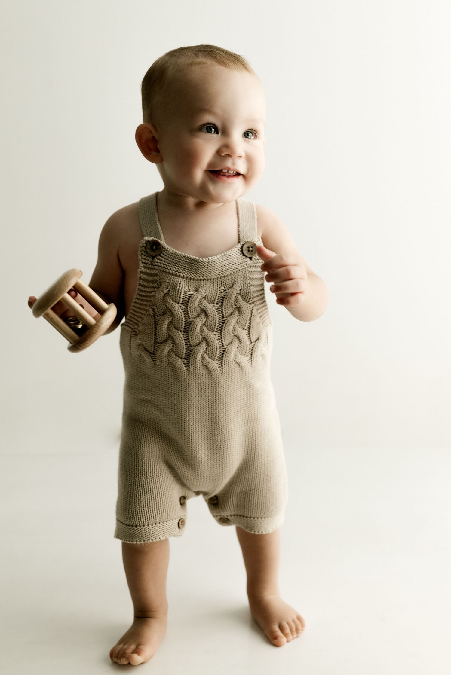 A toddler wearing a knitted beige romper stands barefoot holding a wooden toy.