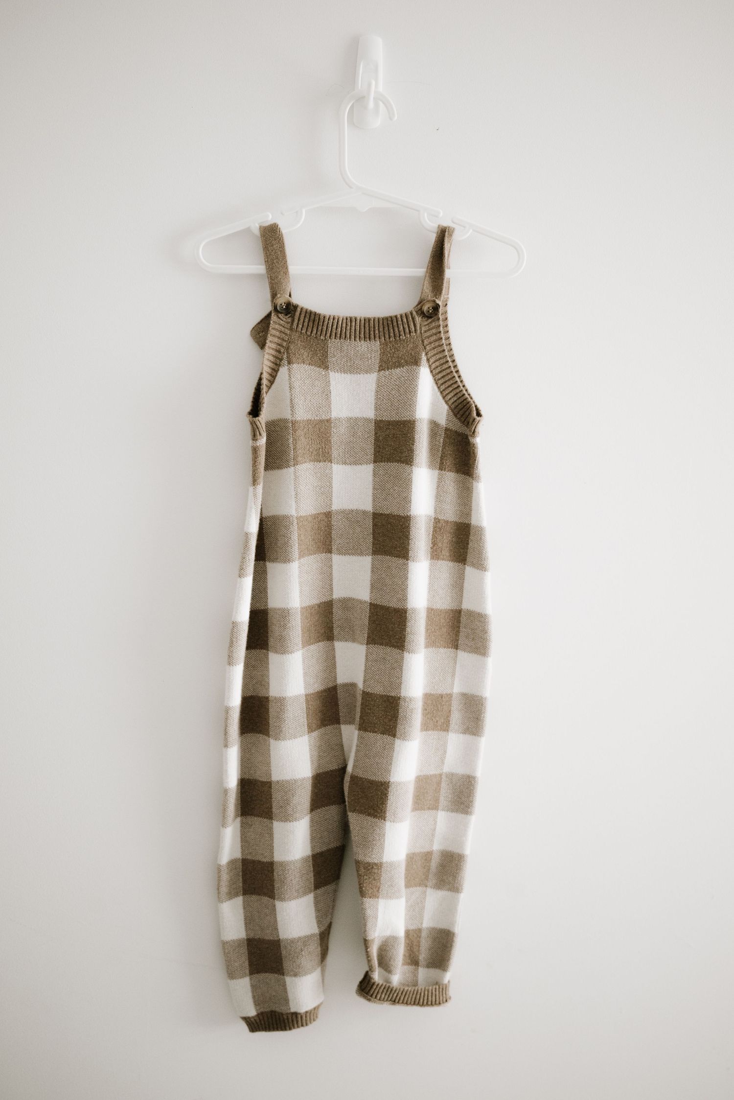 Beige and white checkered knit overalls hanging on white wall.