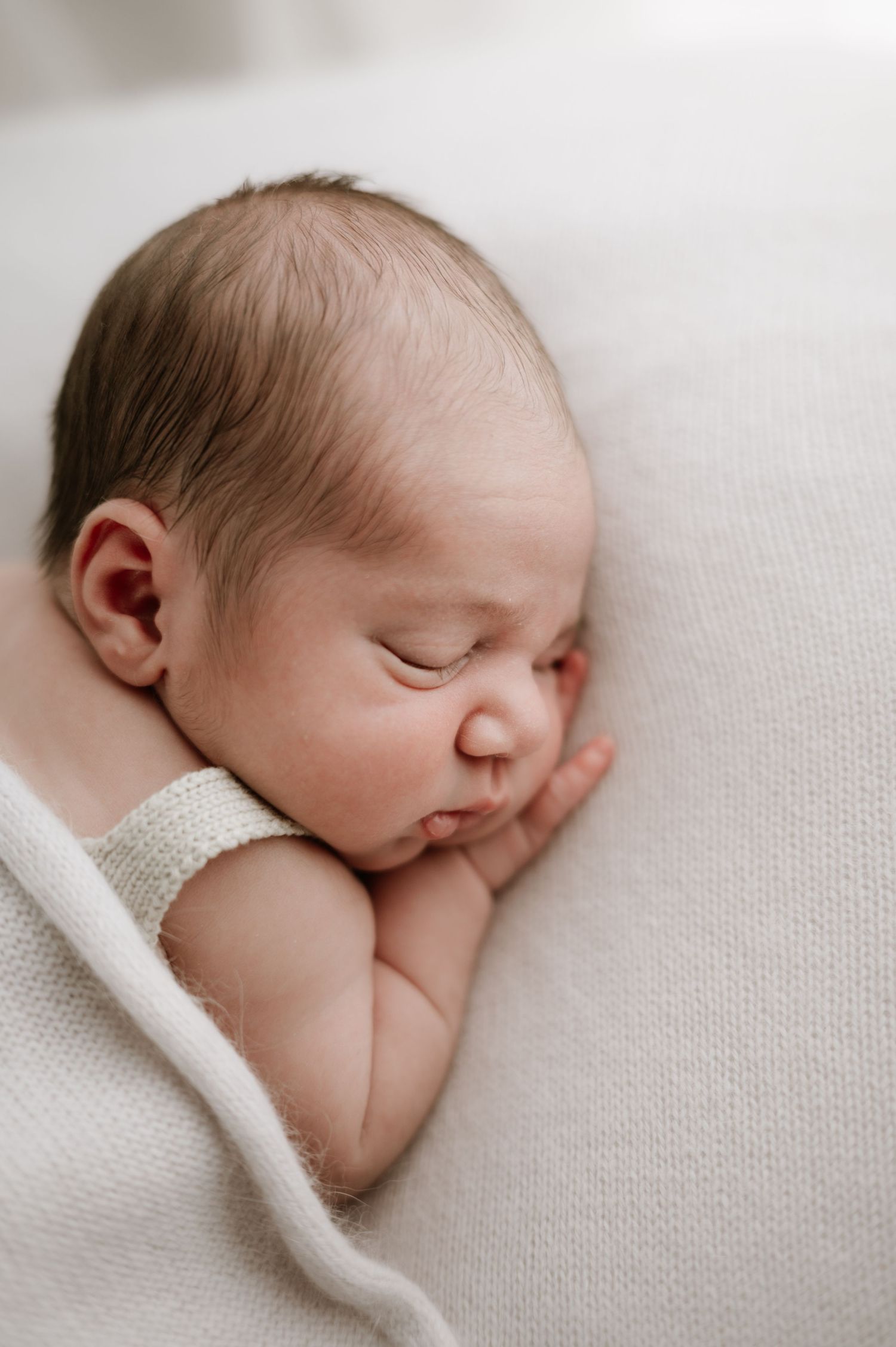 A peaceful sleeping newborn baby wrapped in a soft white blanket against a light colored background.