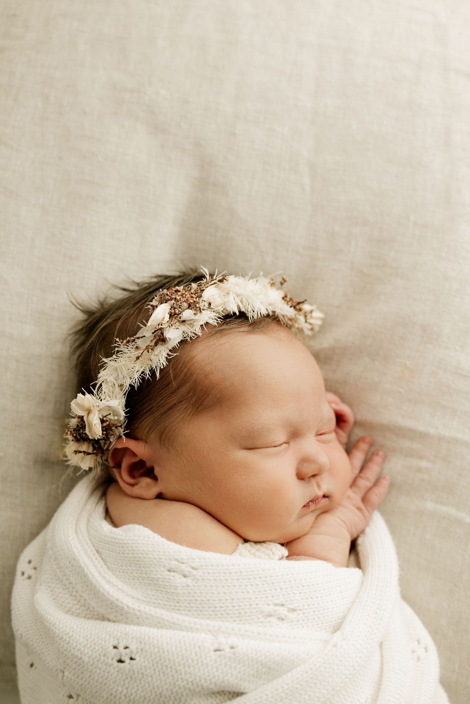 Sleeping newborn wrapped in white blanket wearing delicate floral crown.