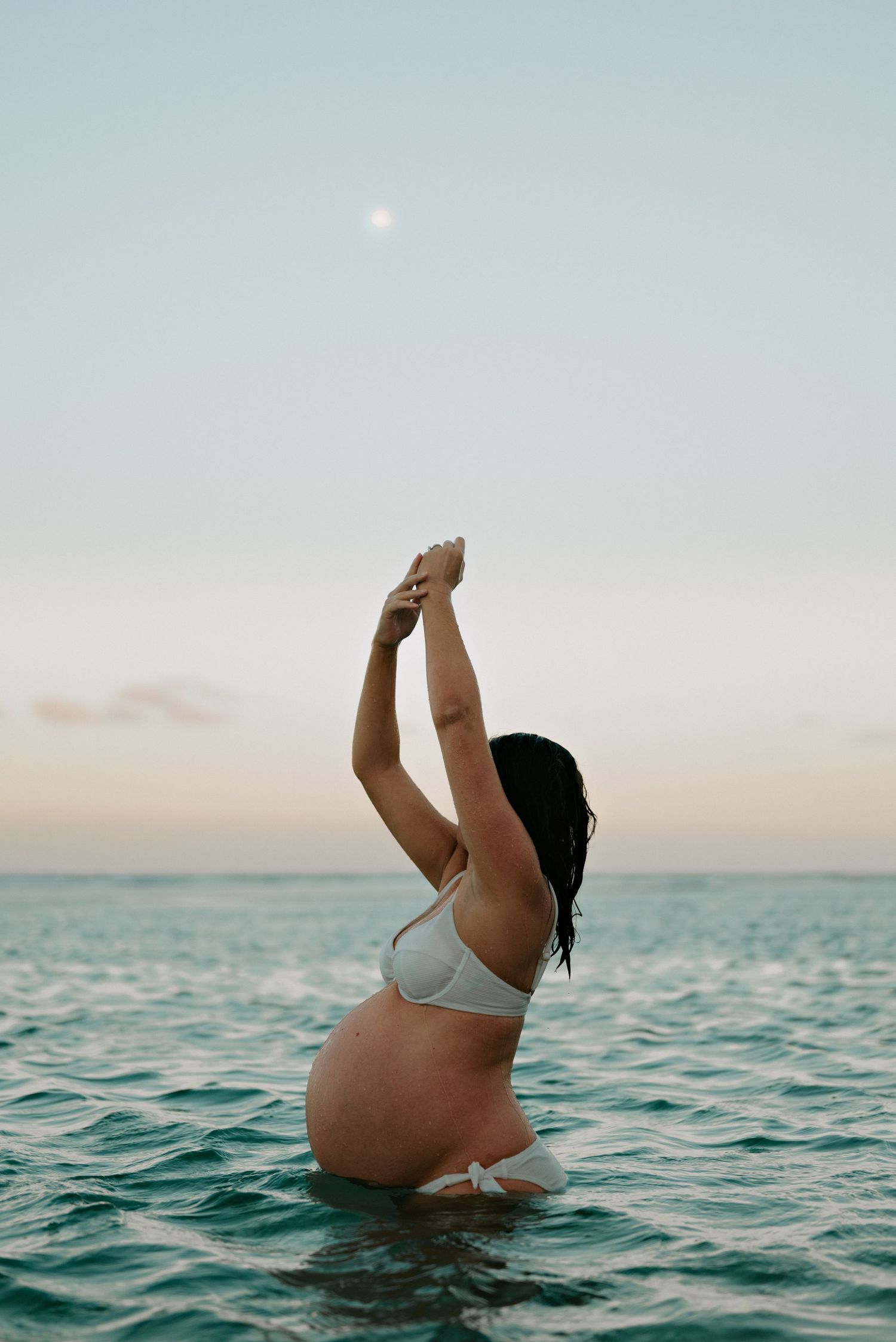 Silhouette of pregnant woman in white bikini raising arms in ocean at sunset.