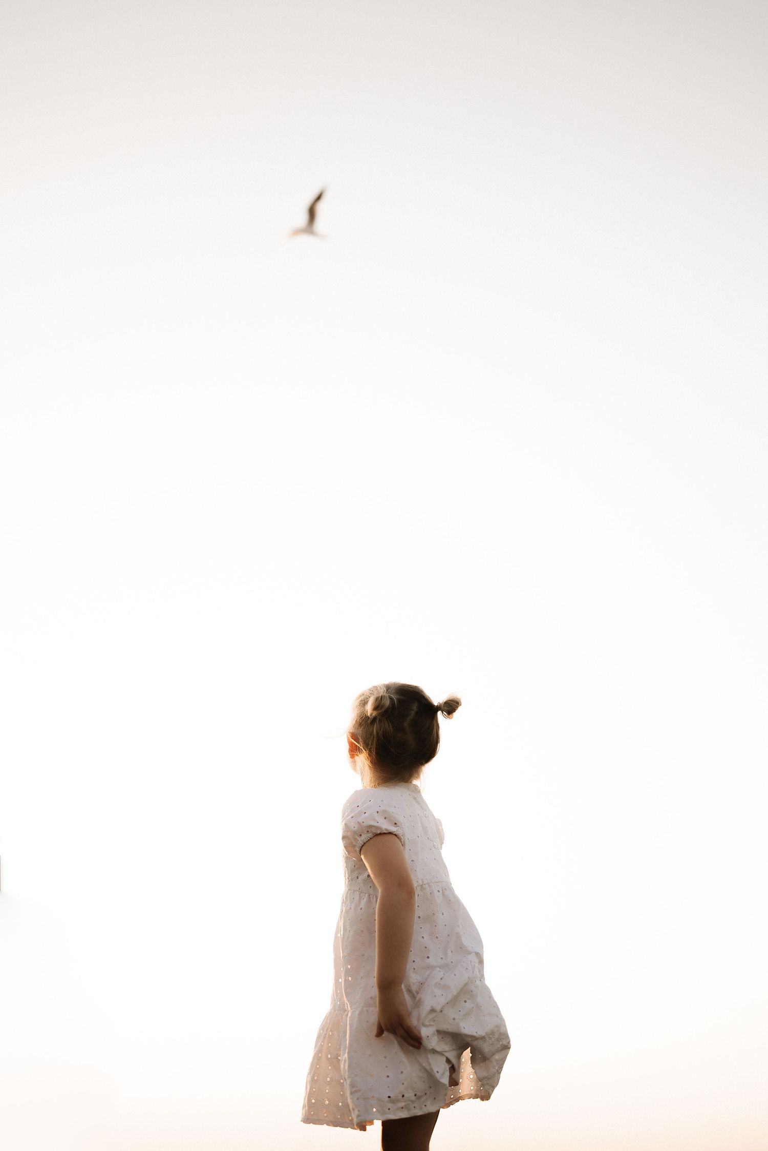 Child in white dress watches bird flying in minimalist bright sky composition.