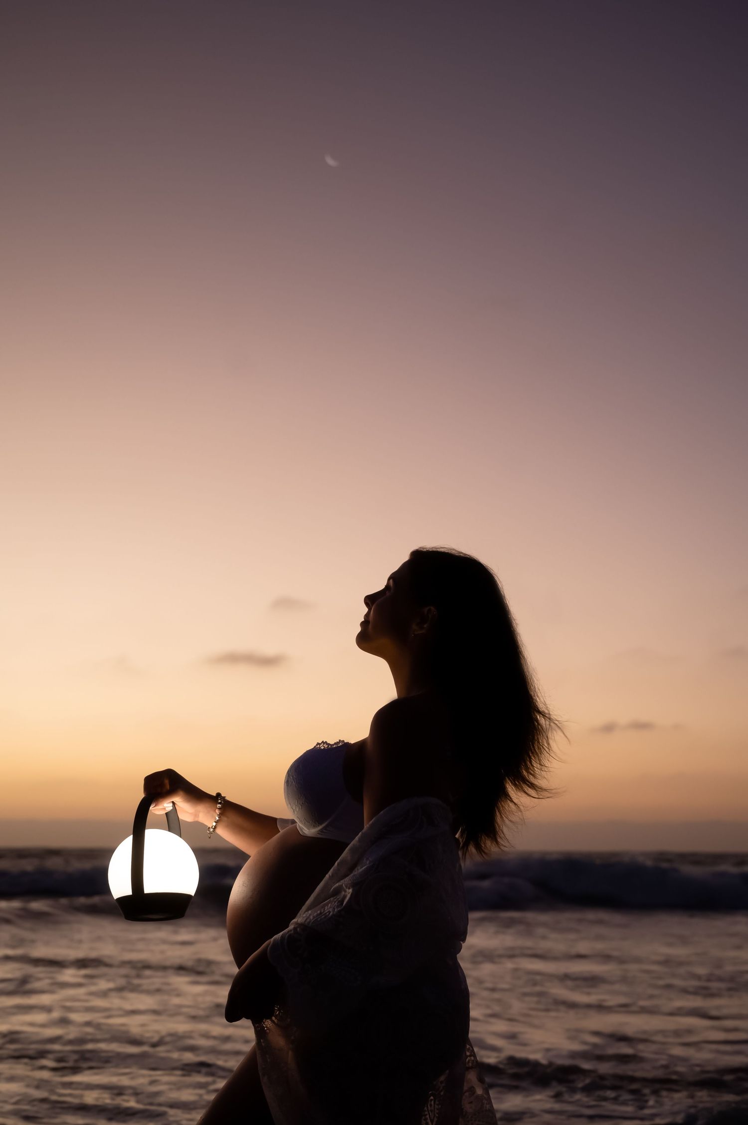 Silhouette of someone holding a lantern by the ocean at sunset with a crescent moon in the dusky sky.