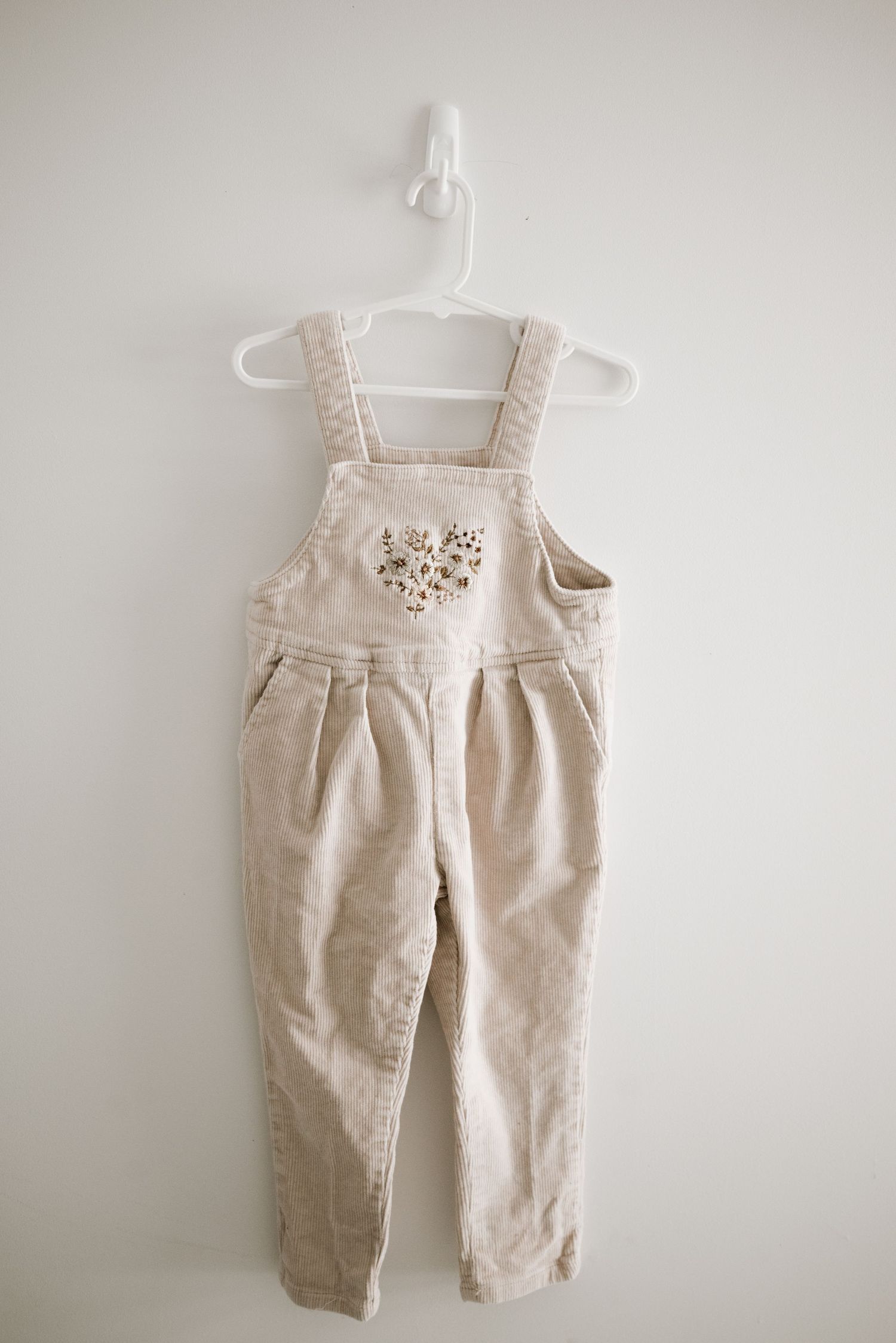 Beige corduroy overalls with heart detail hanging on white plastic hanger against light wall.