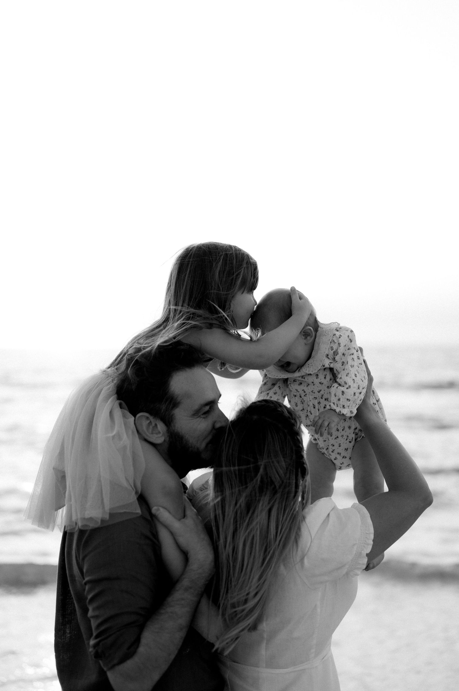 Black and white beach portrait captures loving family embrace against ocean backdrop.