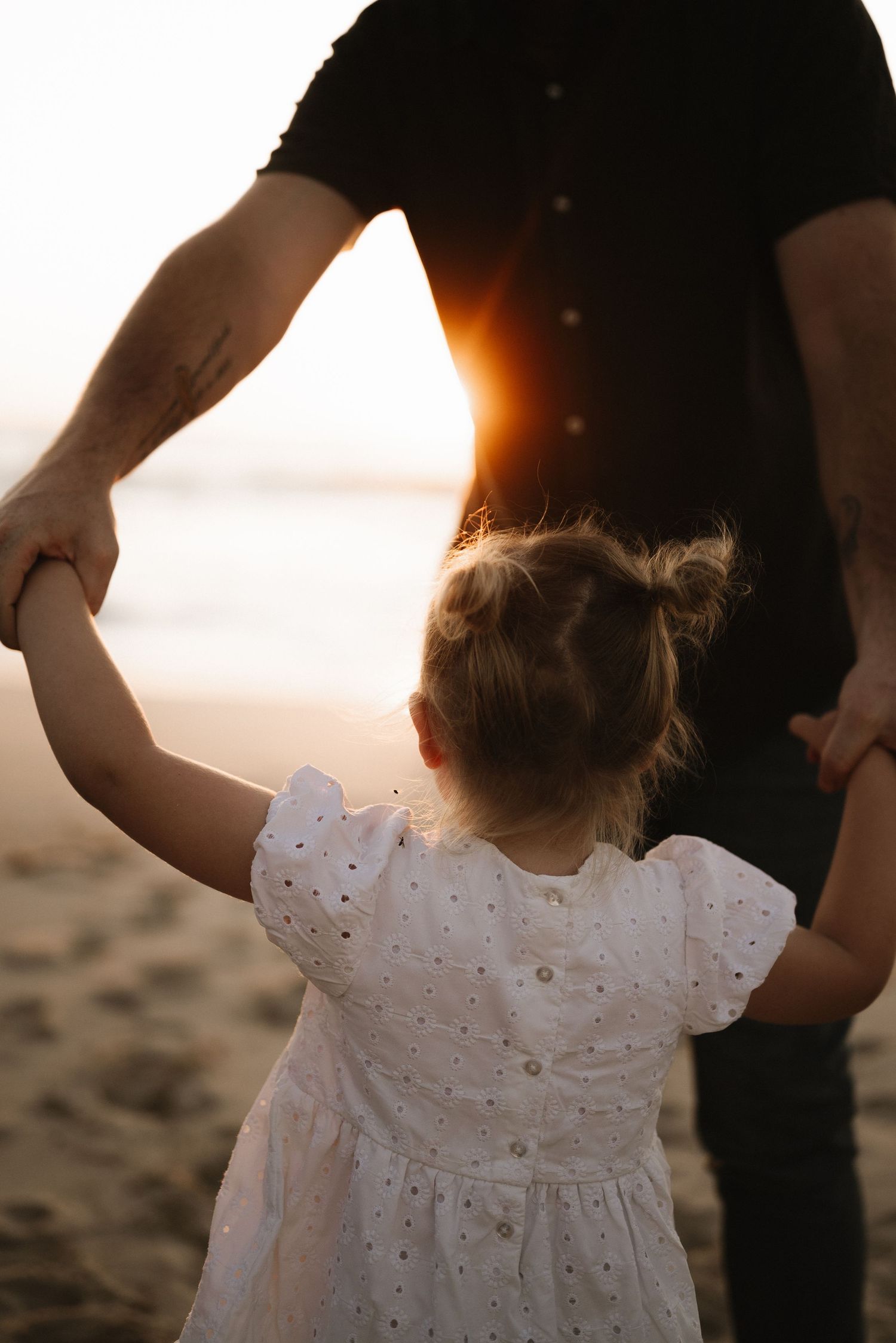 Silhouette of parent and child in white dress holding hands on beach at golden hour.