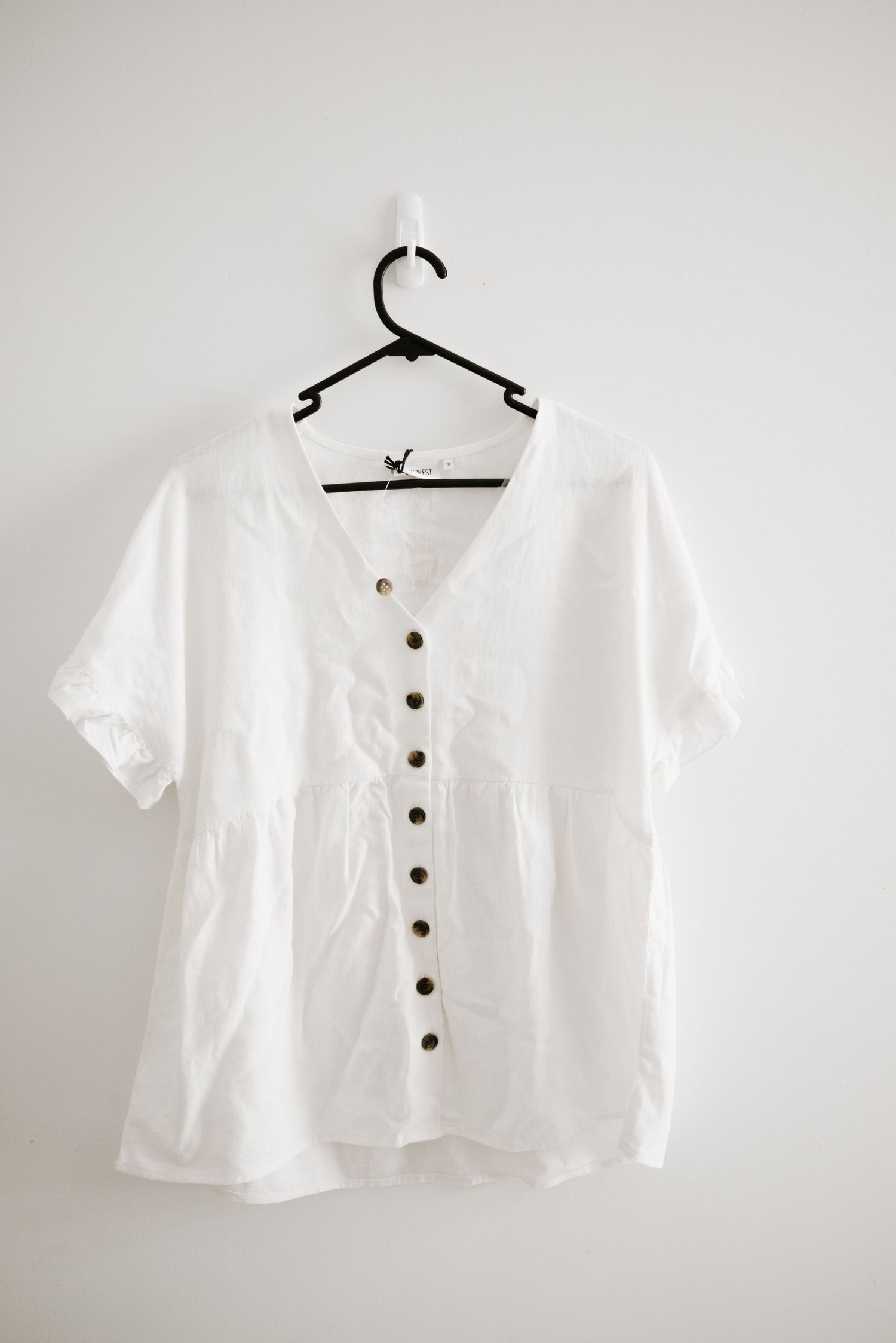 White cotton button-up blouse with short sleeves hanging on black plastic hanger.