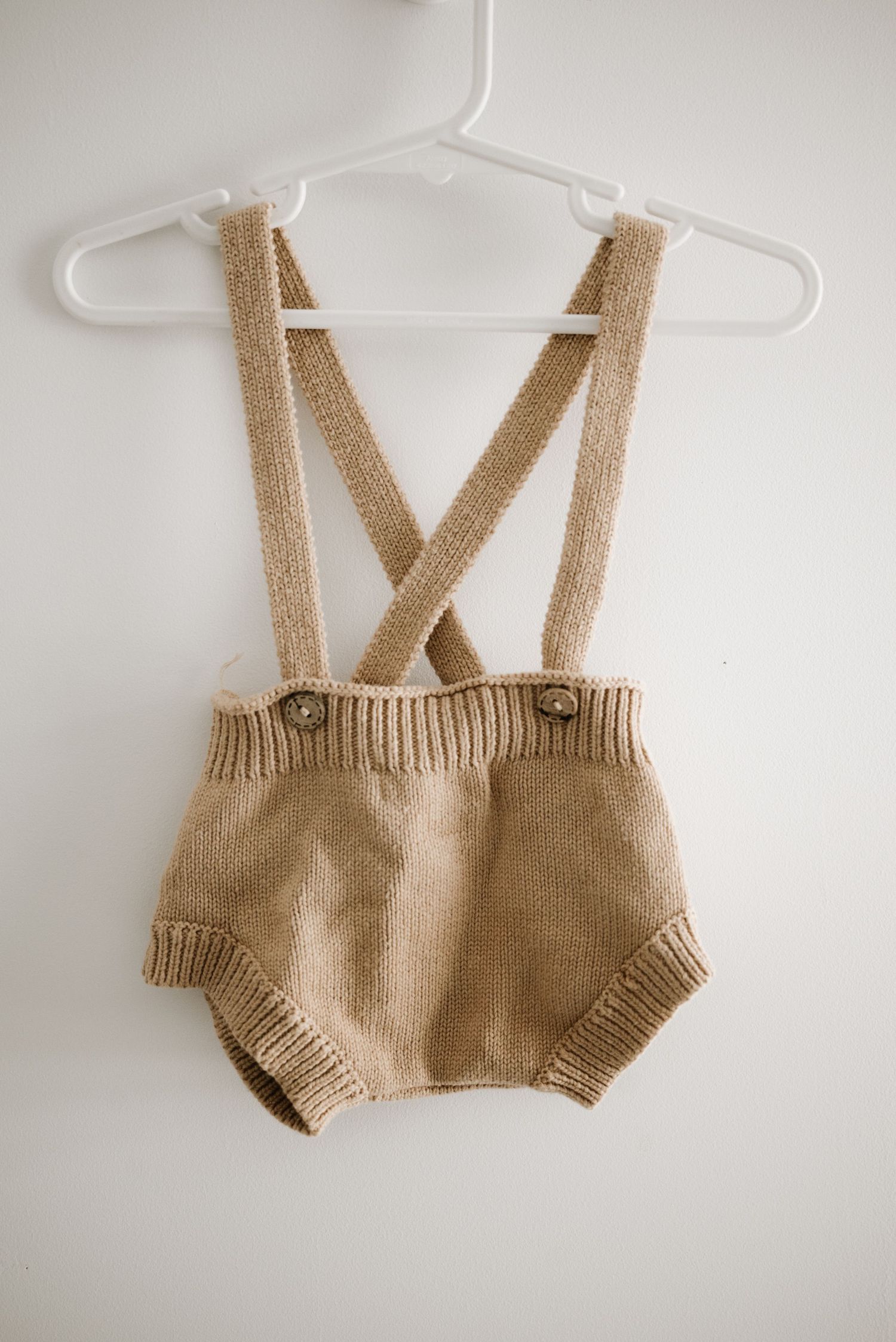 Beige knit baby romper with crossback suspenders hanging on white hanger.