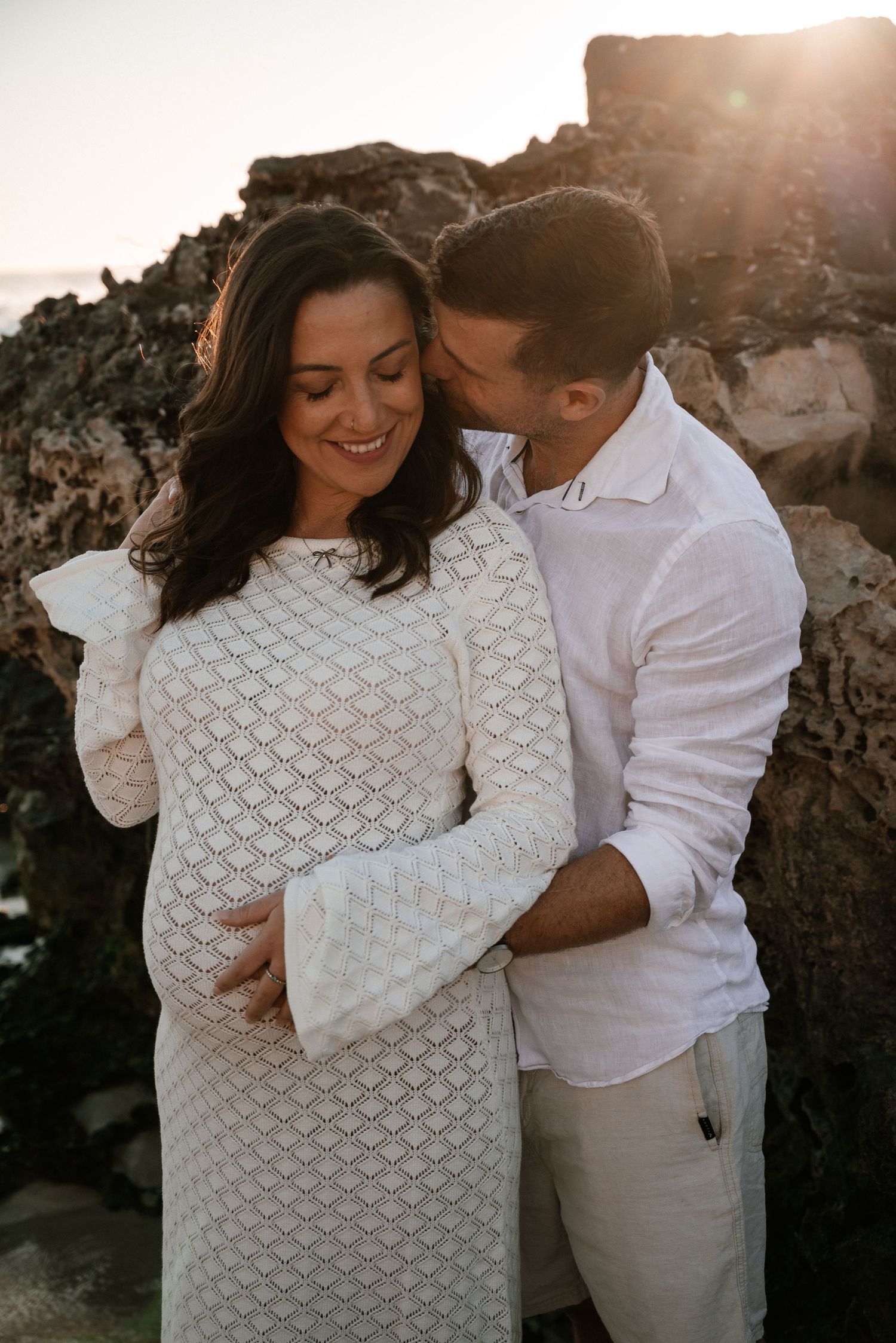 Expecting couple embracing on rocky beach at sunset.