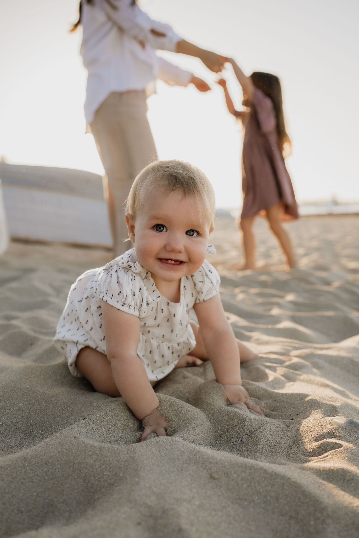 Baby in floral outfit crawling on beach sand while others play in background.
