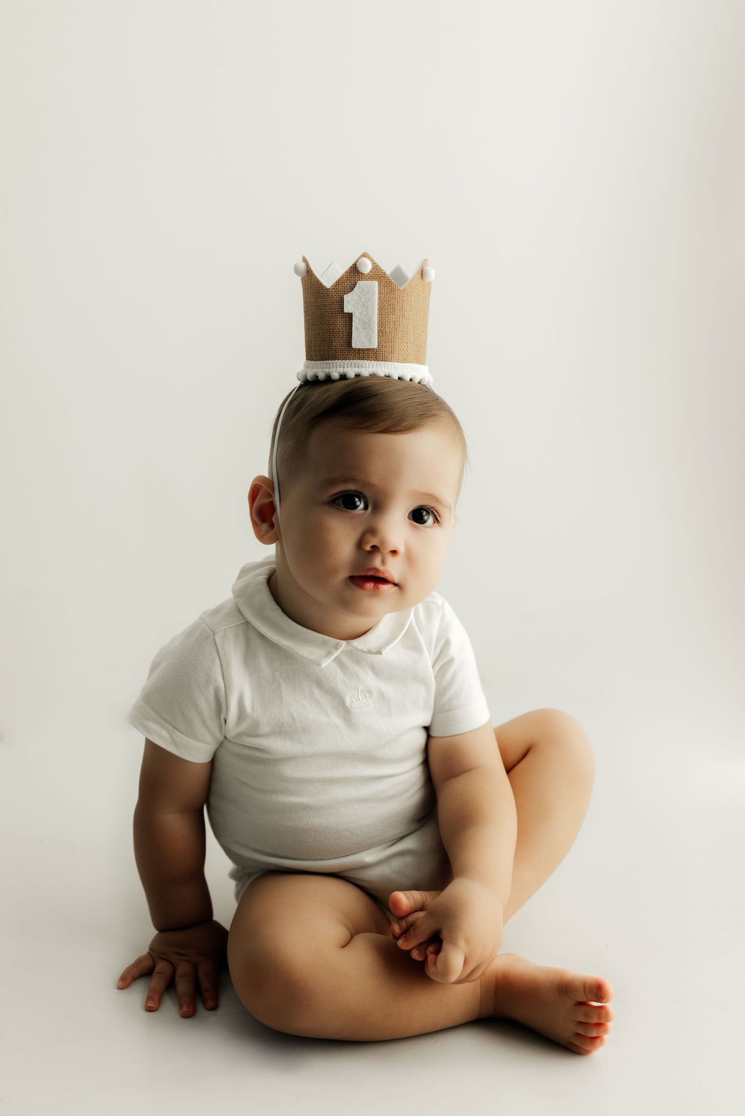 Baby in white onesie wearing gold paper crown sitting on white background.