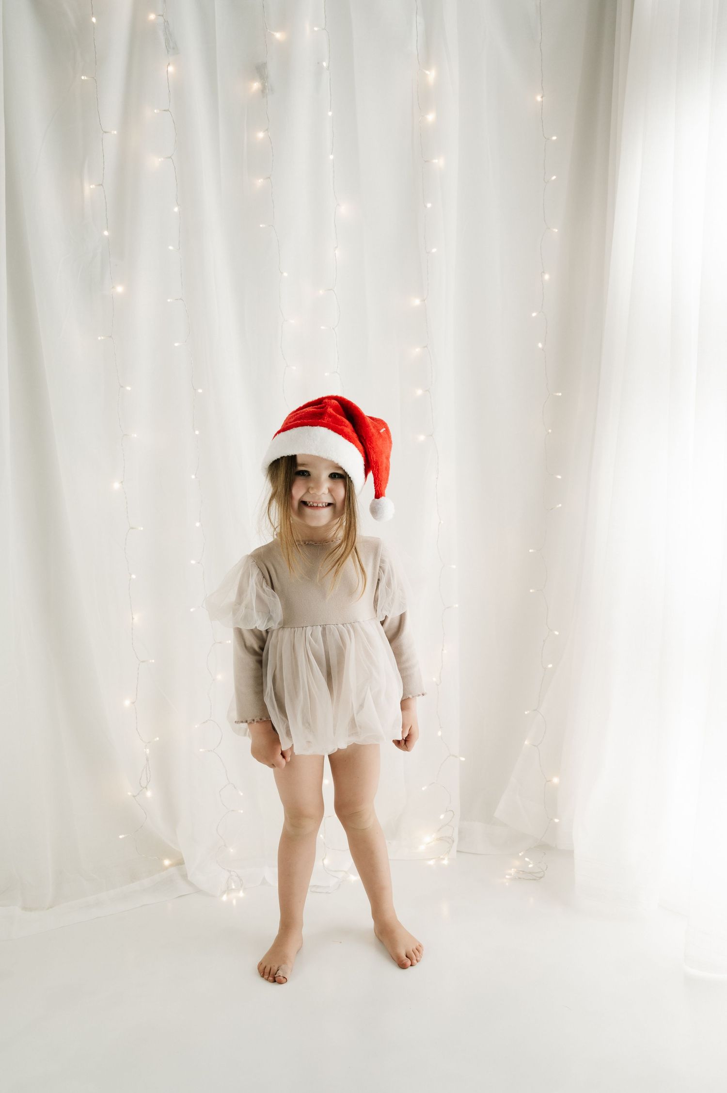 Child in beige dress and Santa hat standing against white backdrop with twinkle lights.