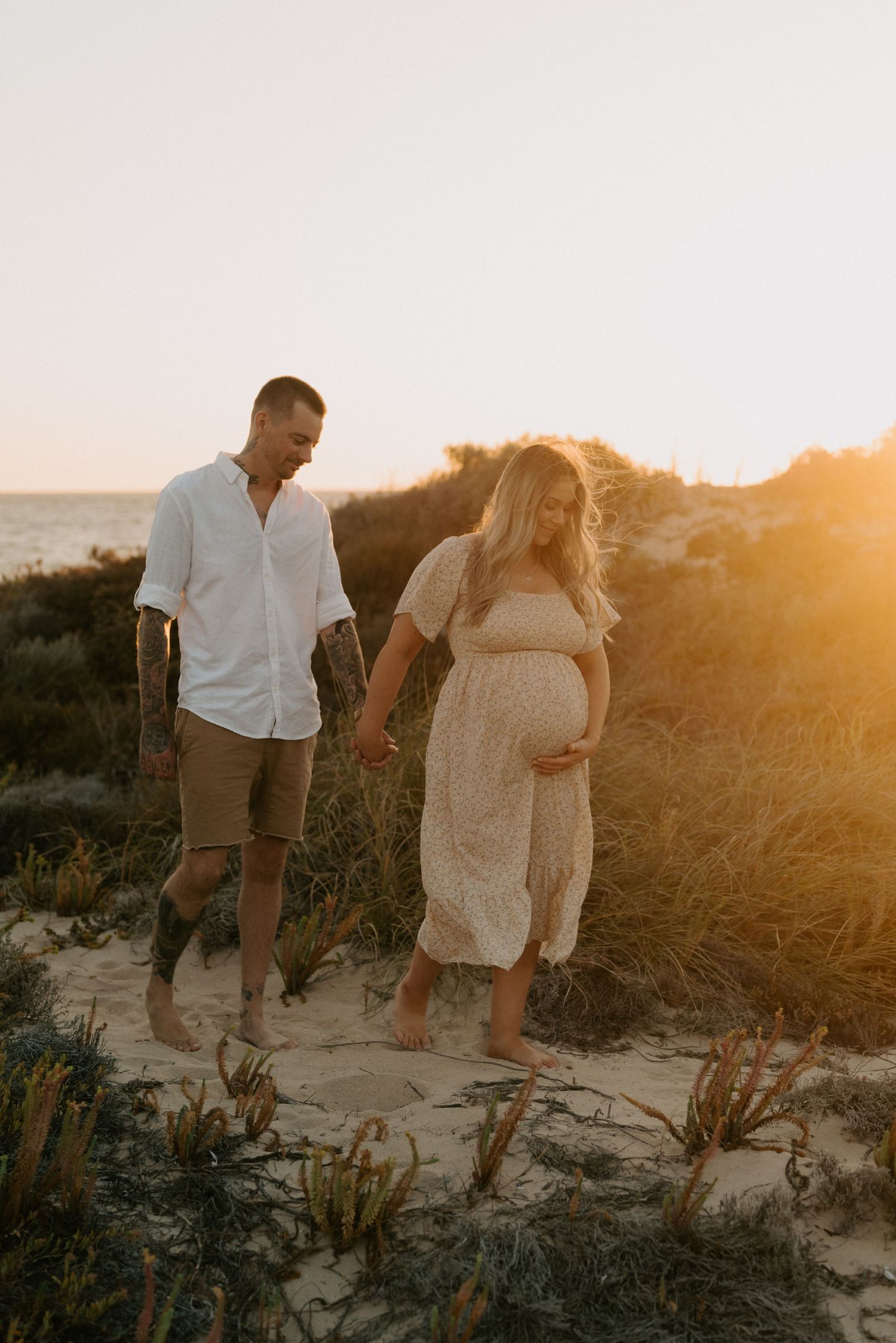 Expectant couple walking on sandy beach at golden hour.