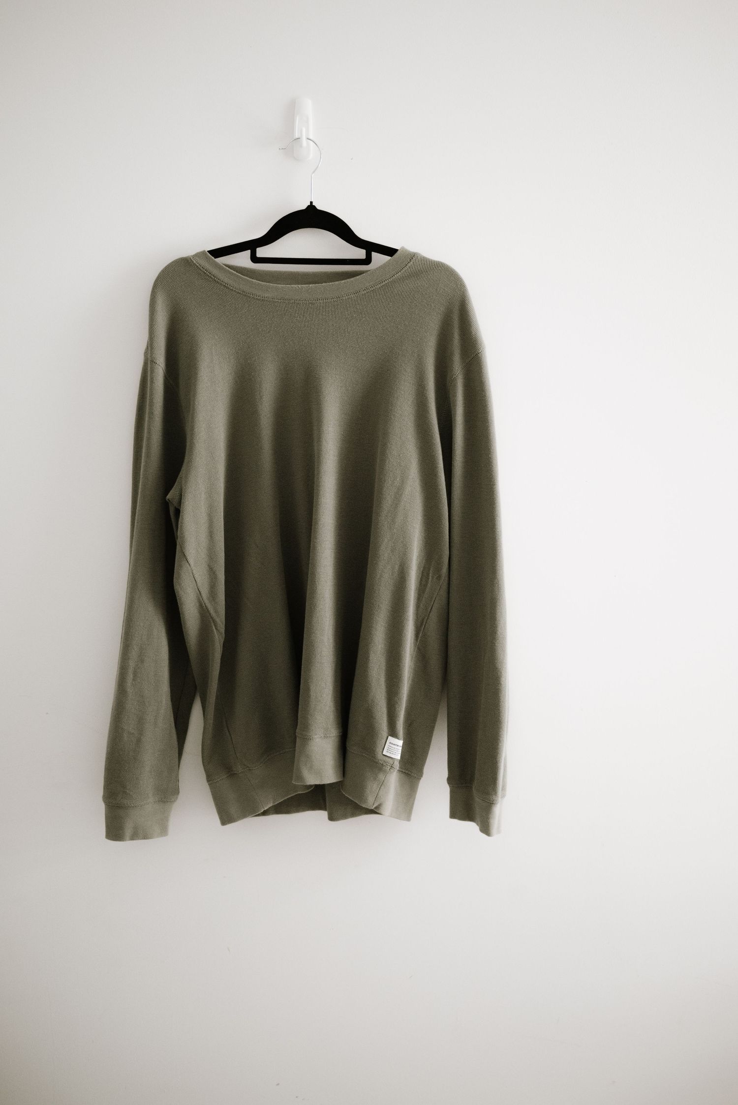 A casual olive green crewneck sweater displayed on a black hanger against a white wall.