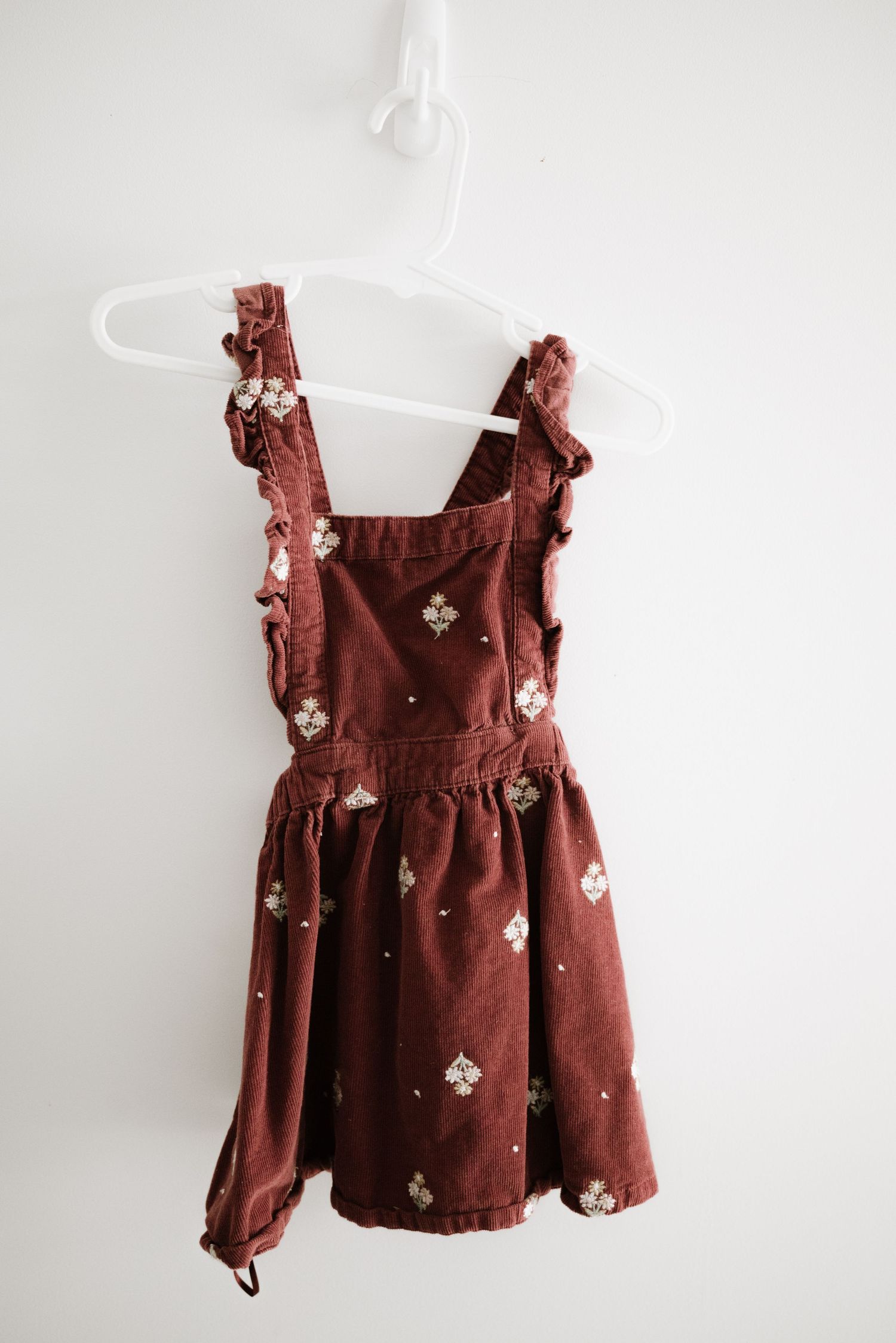 Rust-colored pinafore dress with white floral embroidery and ruffle straps on white hanger.