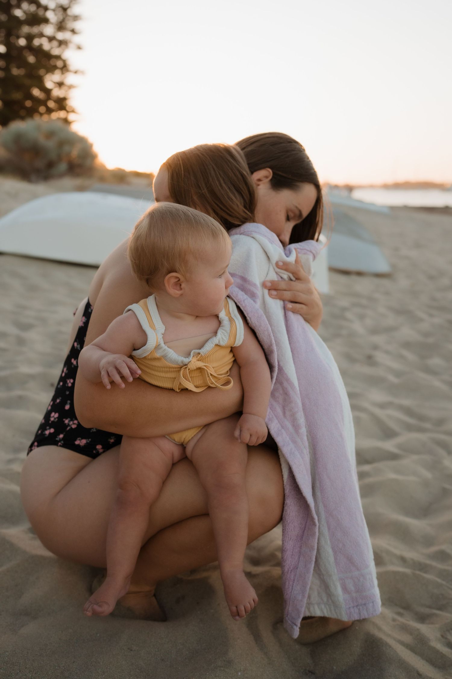Tender beach moment with baby in yellow romper and family embracing.