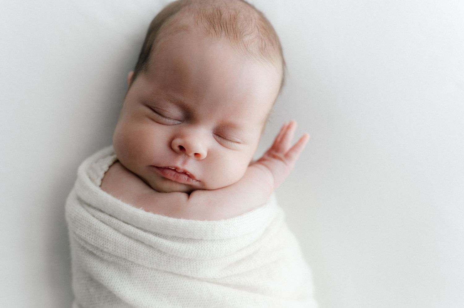 A peaceful sleeping newborn wrapped snugly in a soft white blanket against a light background.