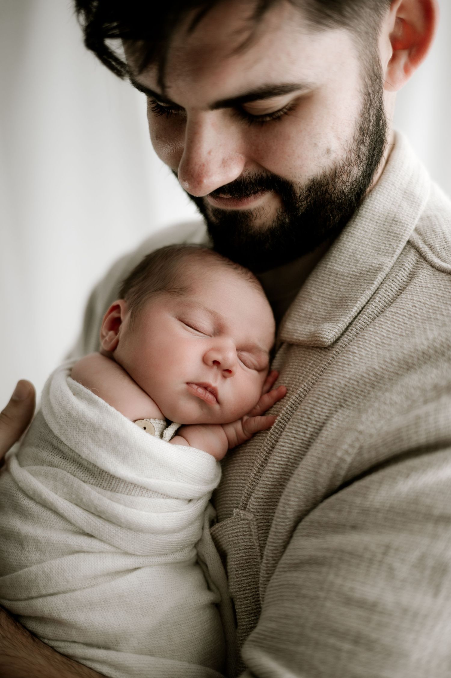 A tender moment between a bearded parent and sleeping newborn baby wrapped in soft white fabric.
