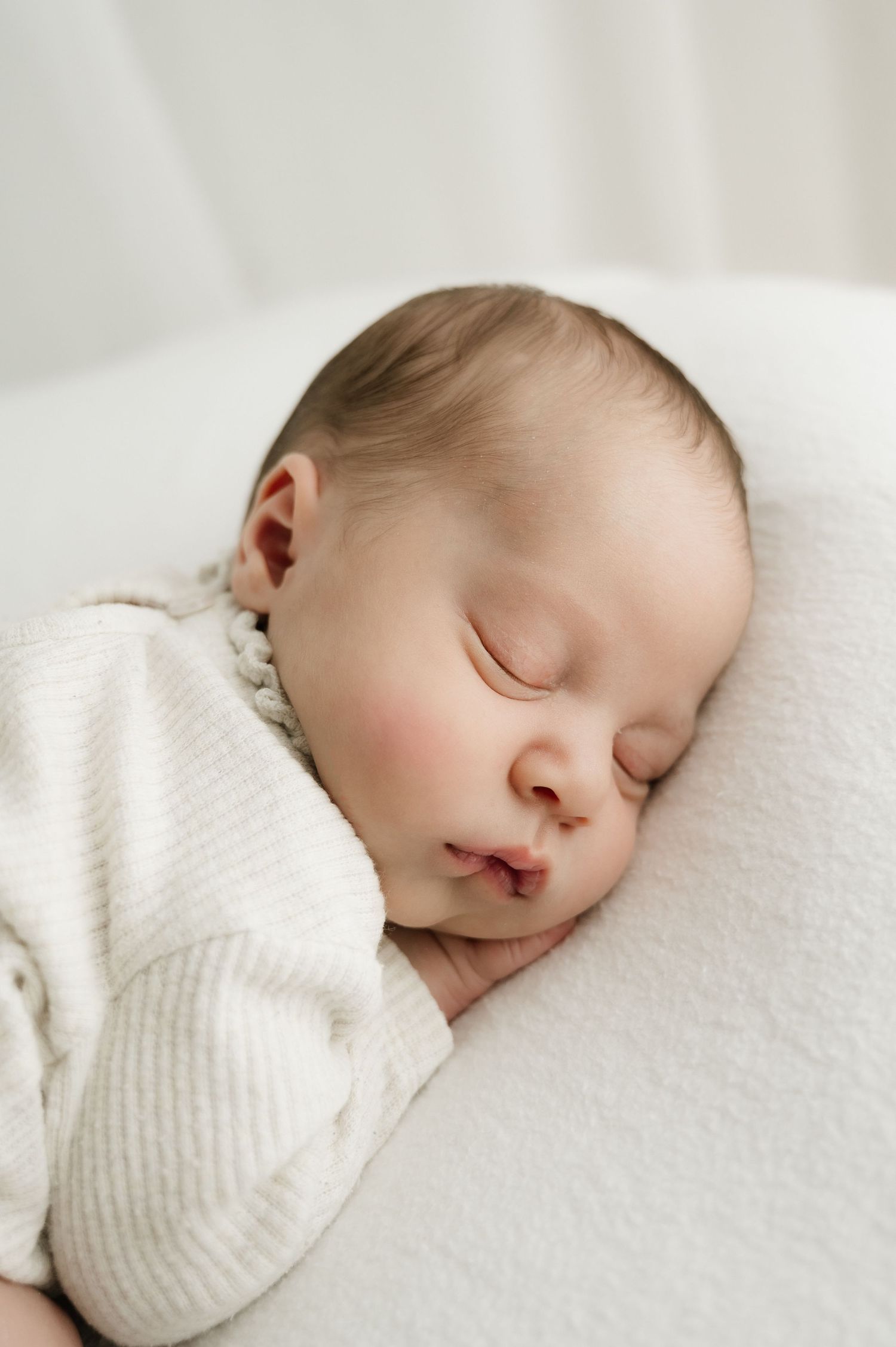 A peaceful sleeping baby in a white sweater rests on soft white bedding with rosy cheeks.