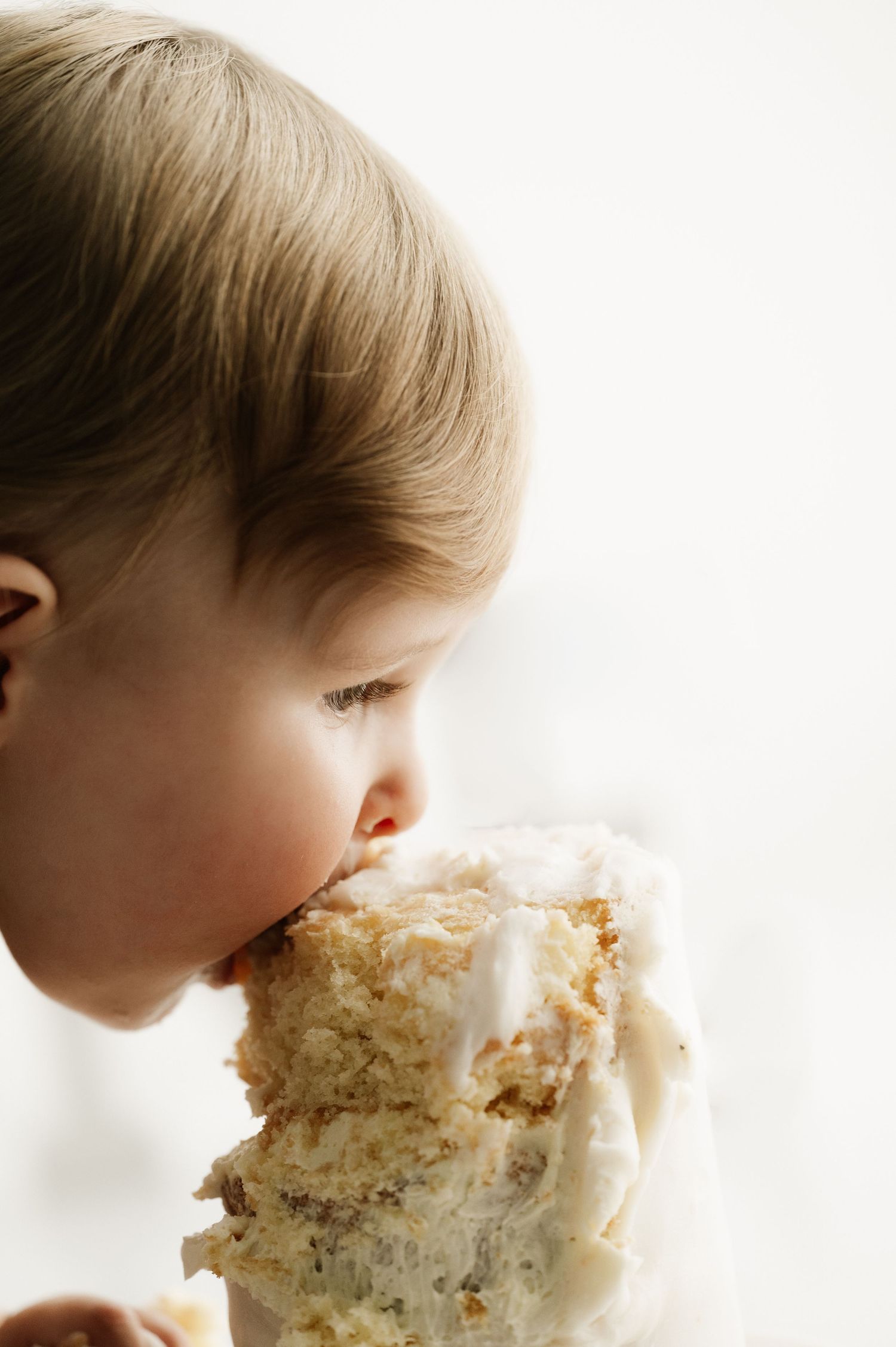 Young child taking a big bite of vanilla frosted cake.