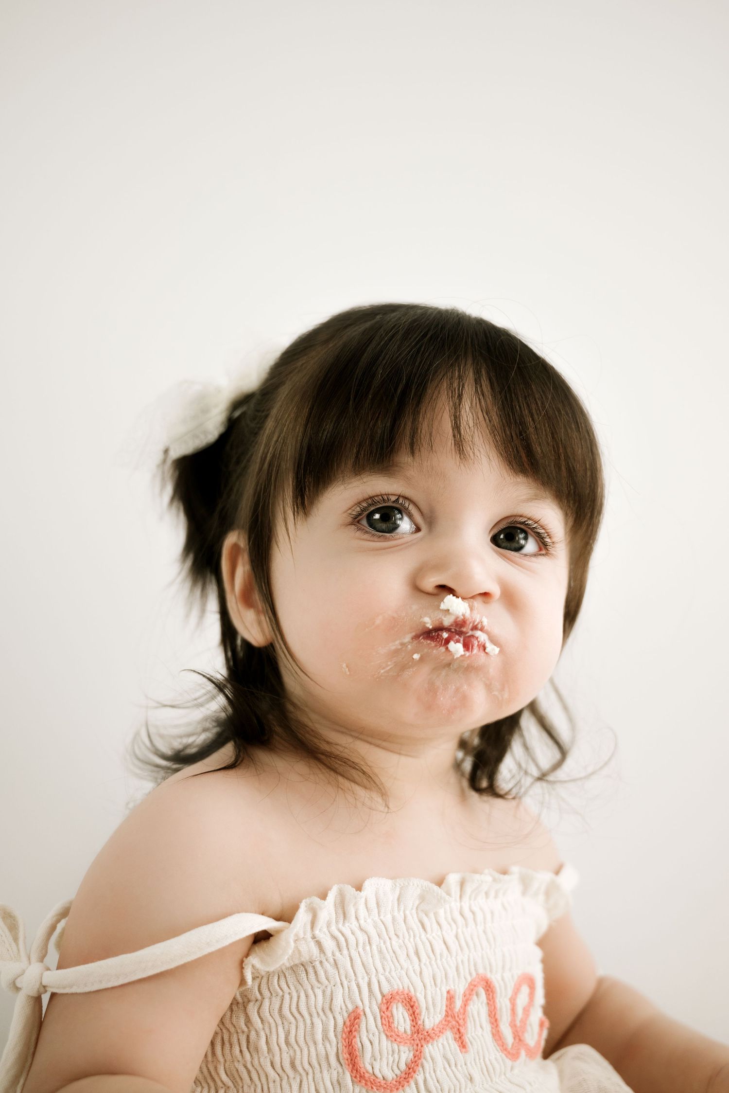 Toddler in a white smocked dress with 'one' written on it looks up with messy face after cake smash photo session.