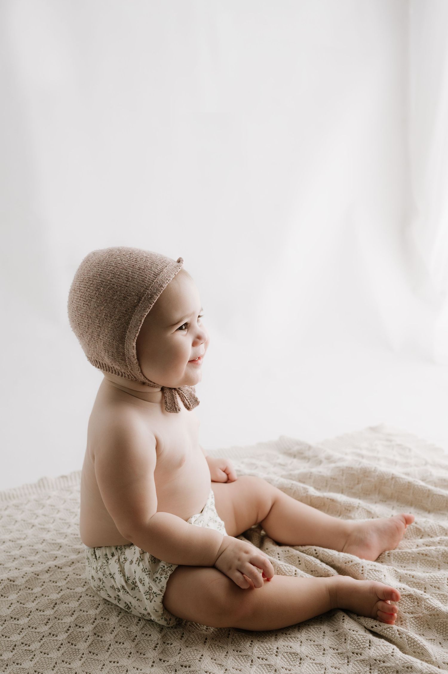 A baby wearing a brown bonnet sits on a textured blanket in a minimalist portrait setting.