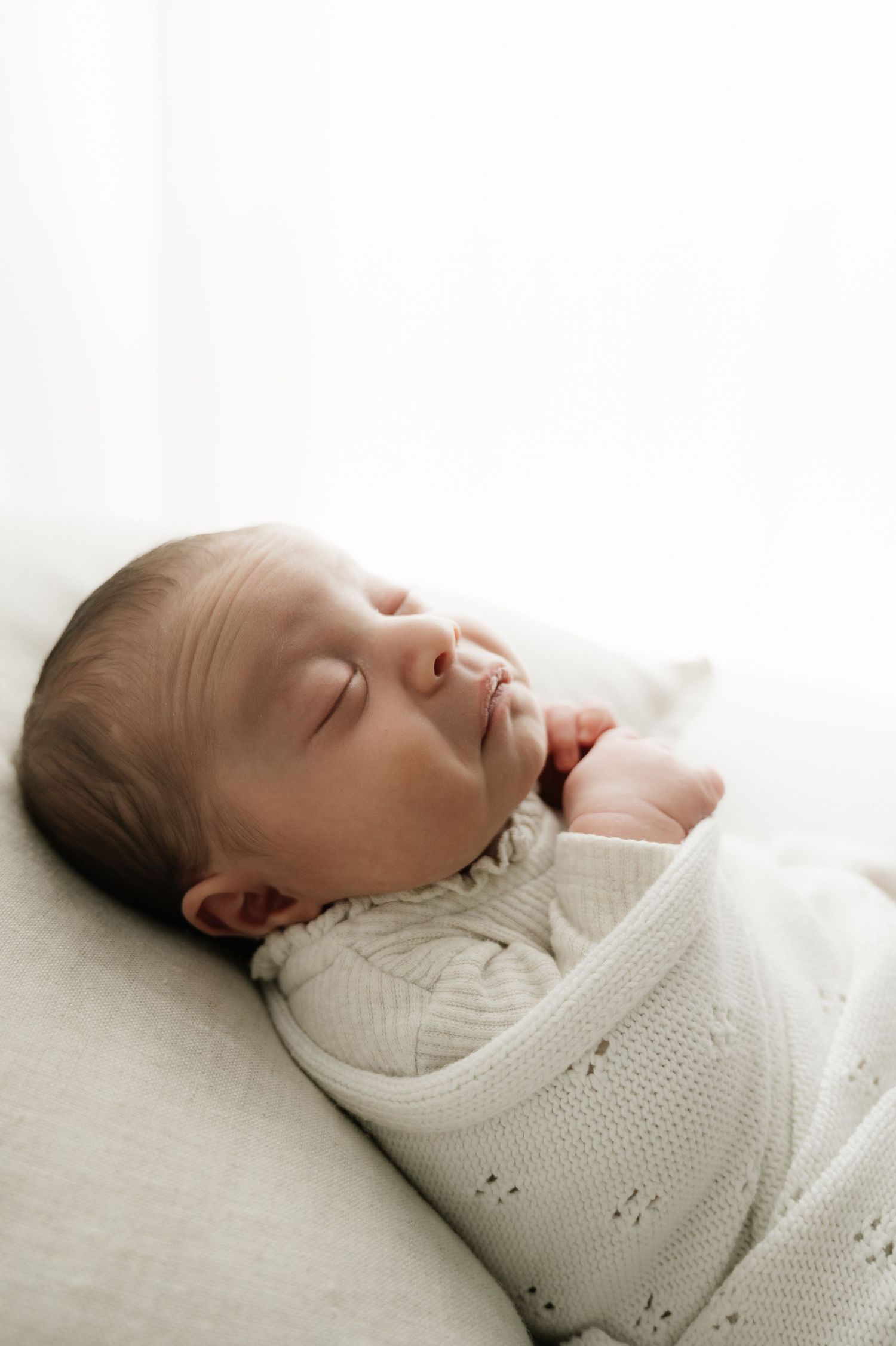 A peacefully sleeping baby wearing a white knit sweater lies on a light-colored cushion in soft lighting.
