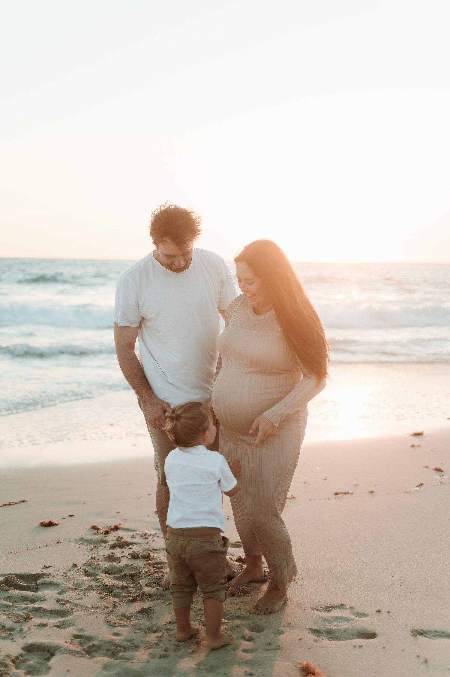 Young couple shares tender moment with their toddler on sunset beach as waves crash behind them.