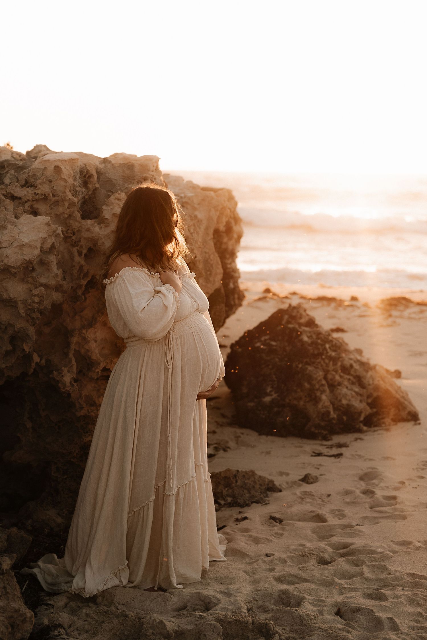 Expectant mother in flowing white dress poses near beach rocks at golden hour.