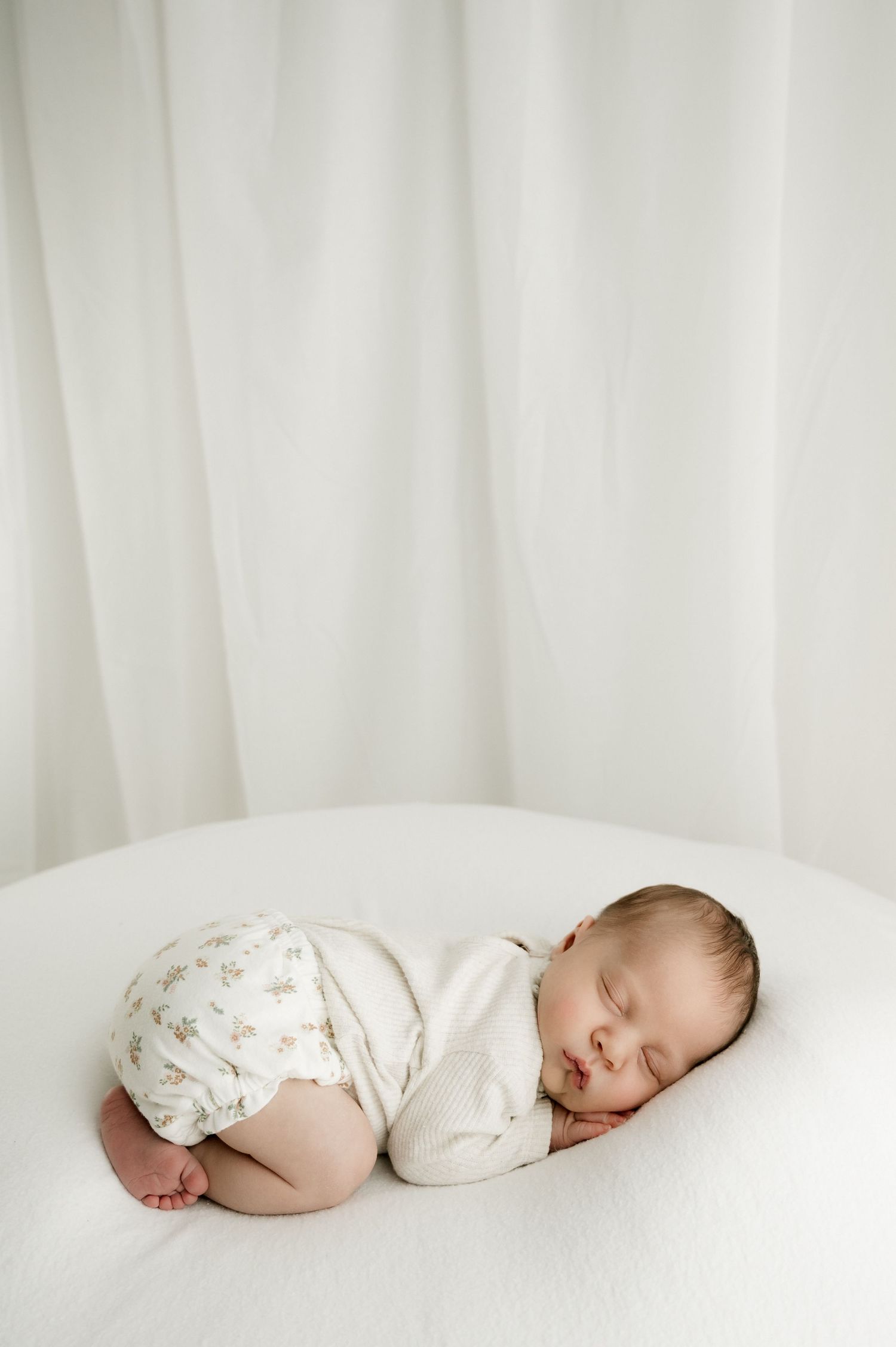 Peaceful sleeping baby in white onesie and floral pants rests on soft white surface against light backdrop.