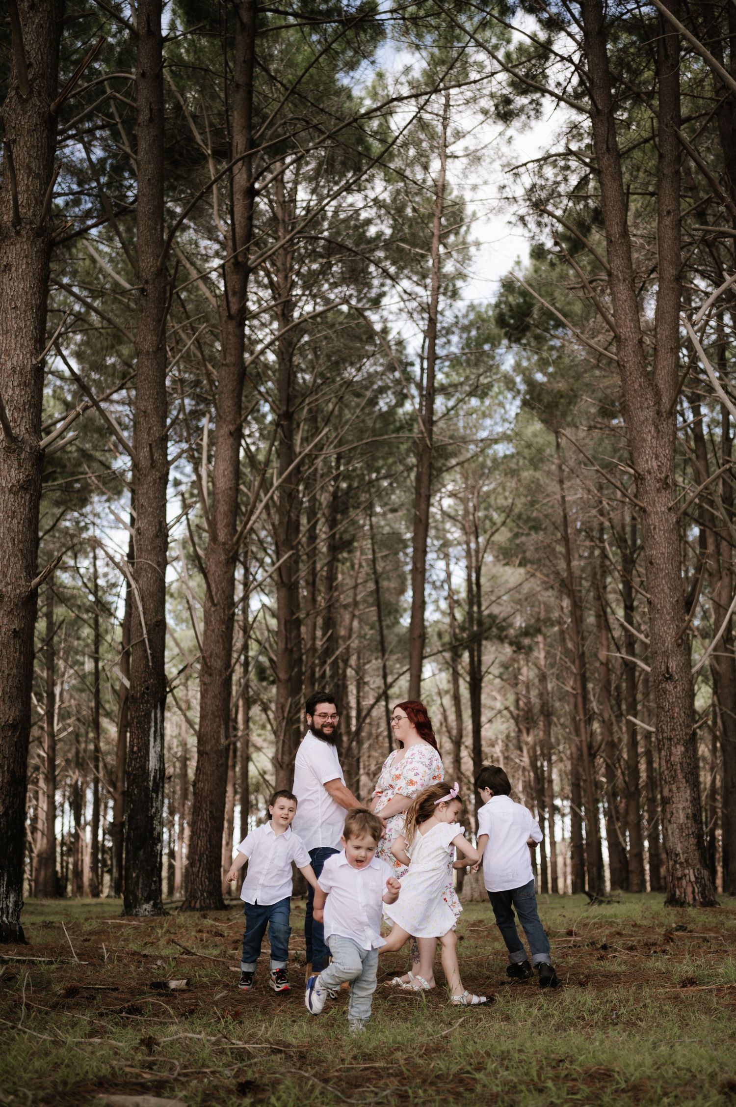 A group wearing coordinated white outfits poses together in a serene pine forest setting during golden hour.