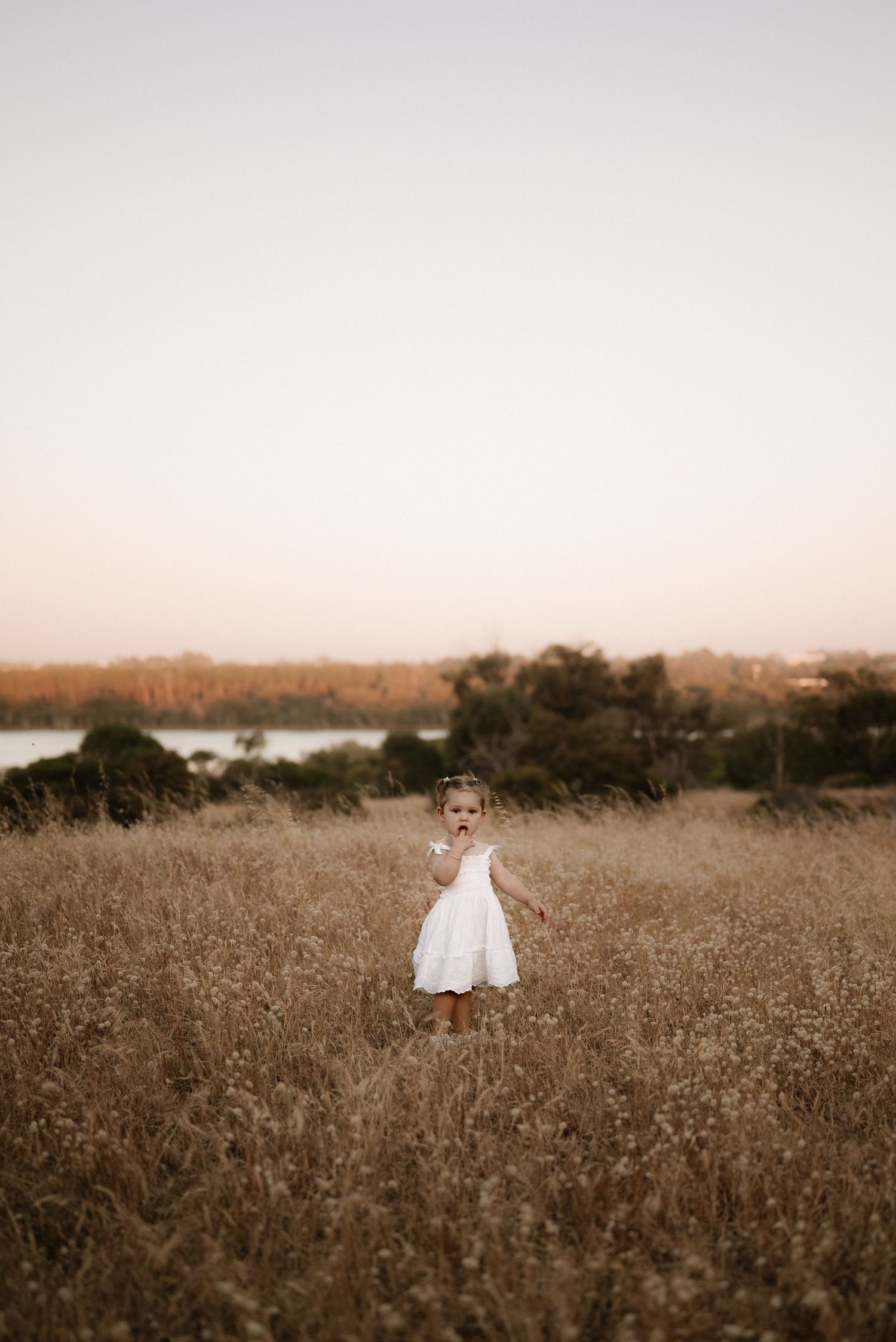 Small child in white dress stands alone in golden field with water view.