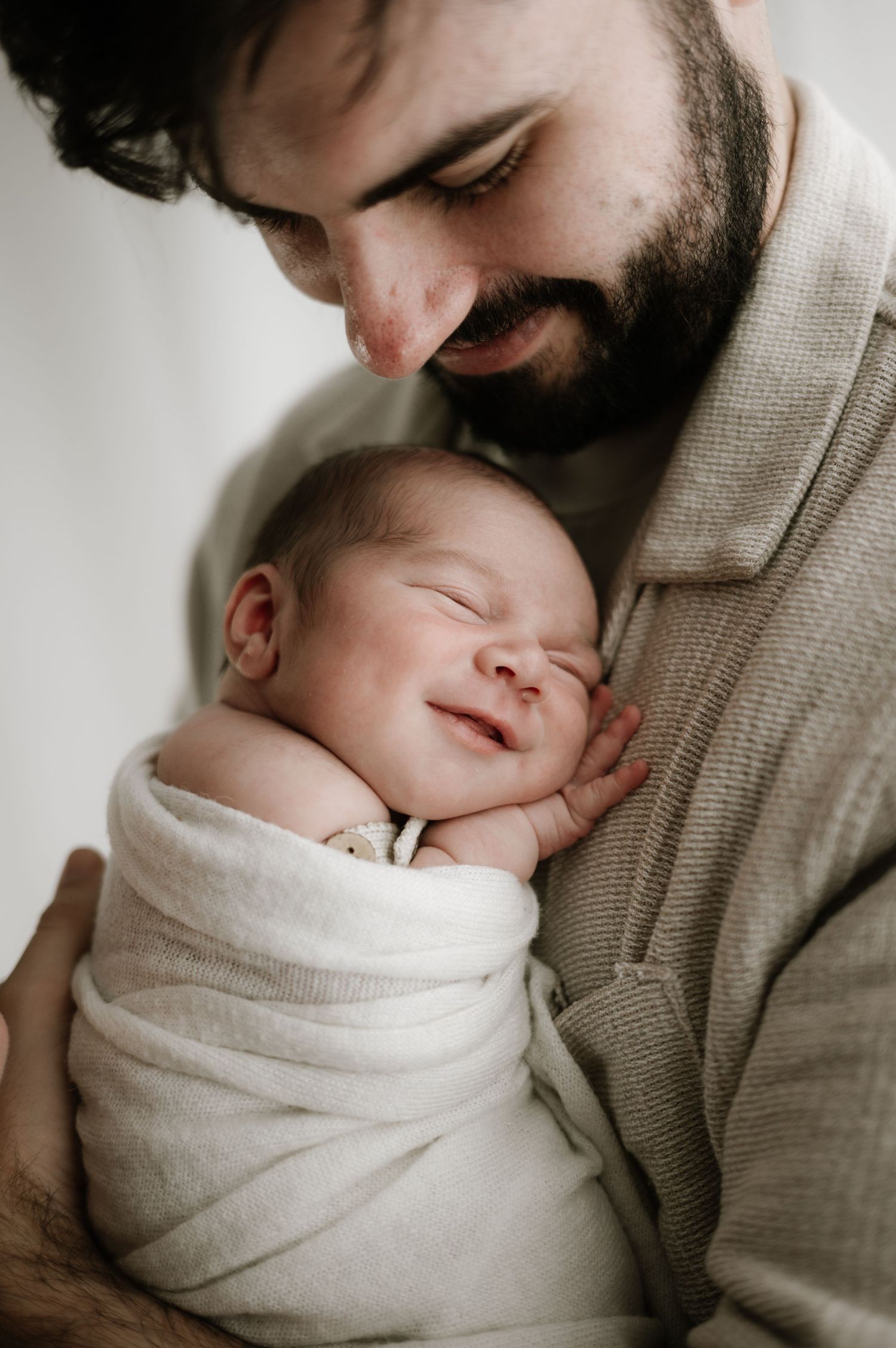 A tender moment between a bearded parent holding a sleeping newborn baby swaddled in soft white fabric.