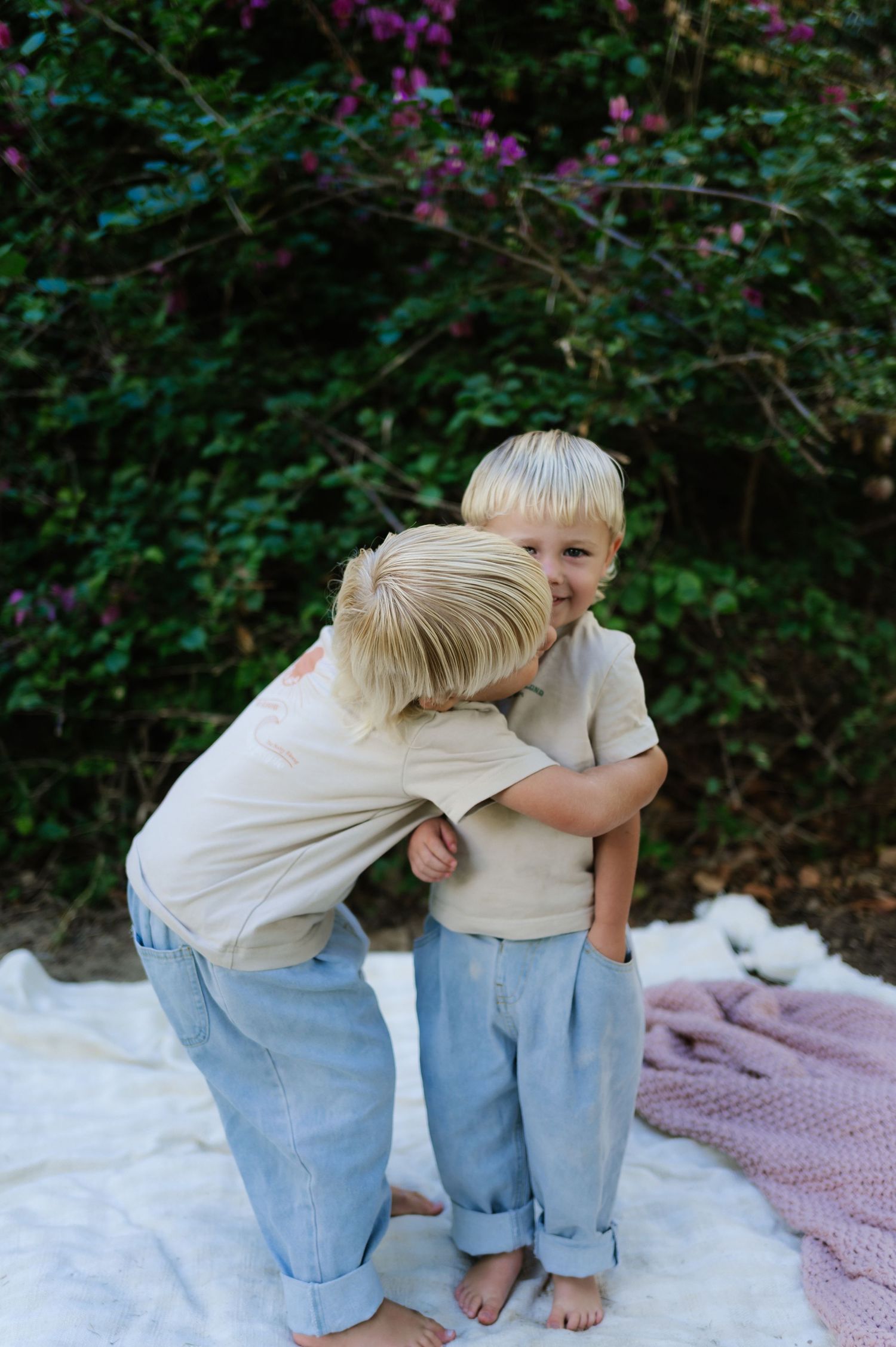 Two children playfully interact while standing on a white blanket with flowering bushes in the background.