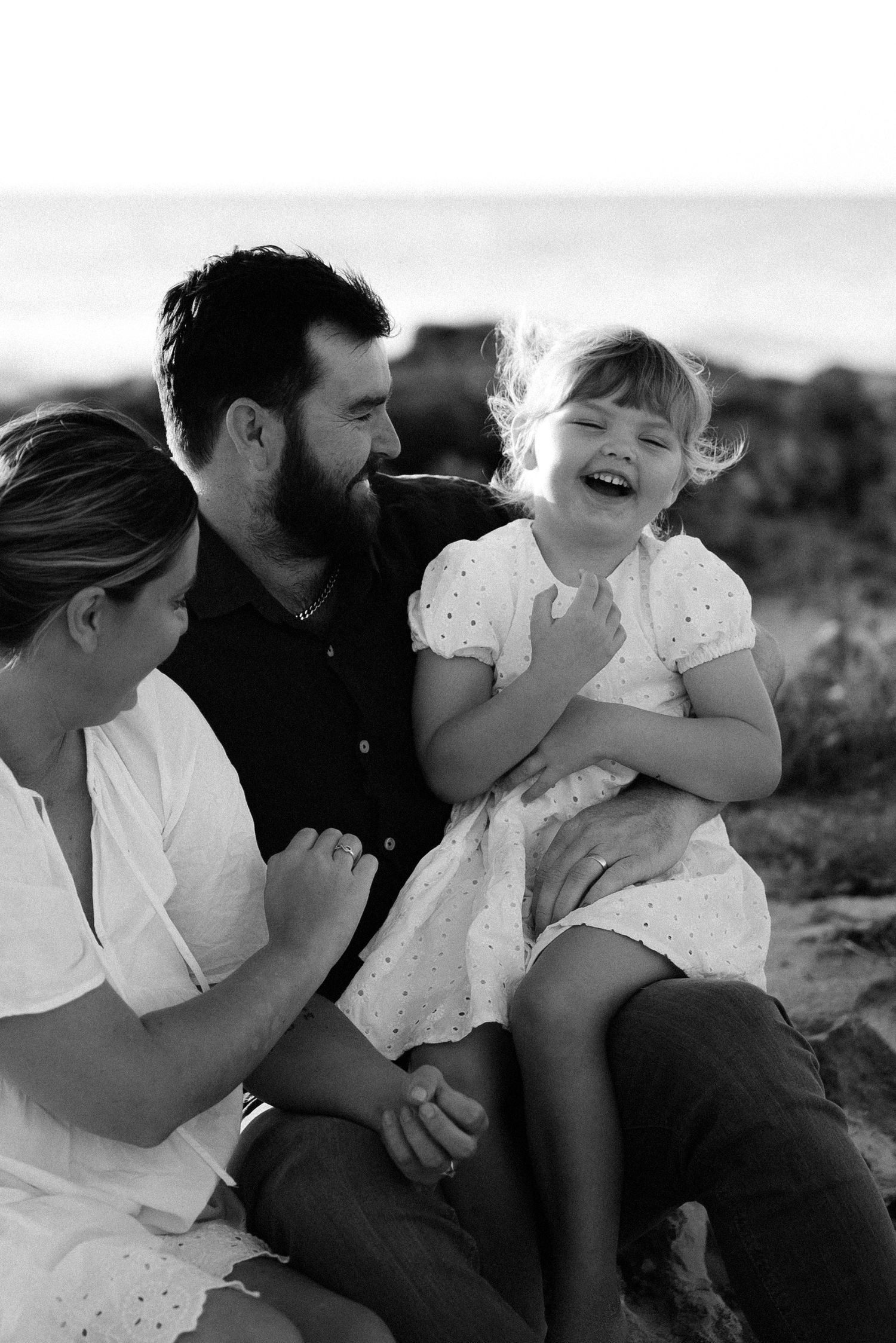 A candid black and white family portrait shows joyful laughter during an outdoor photoshoot.