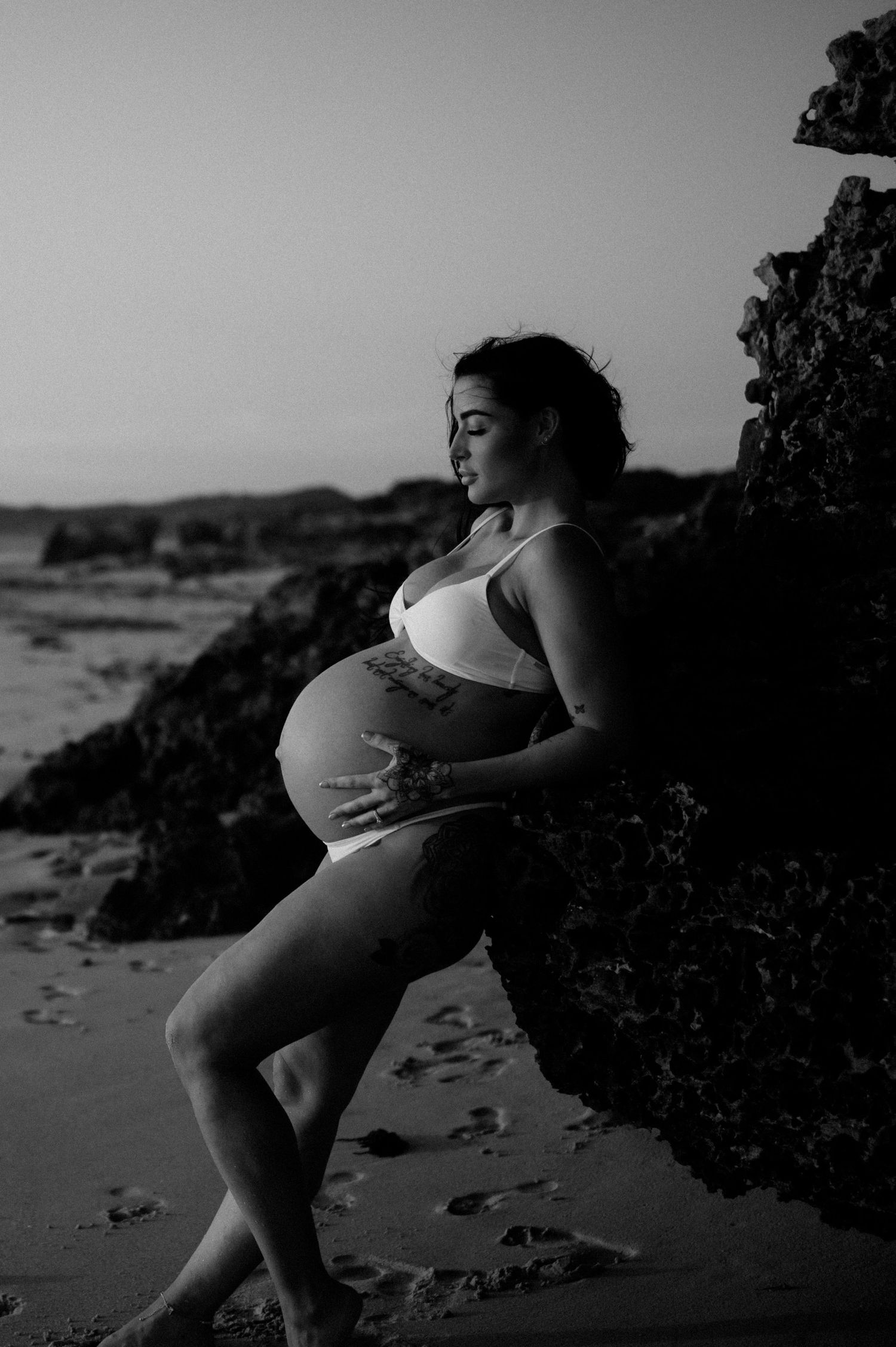 Black and white portrait of pregnant woman posing against beach rocks.
