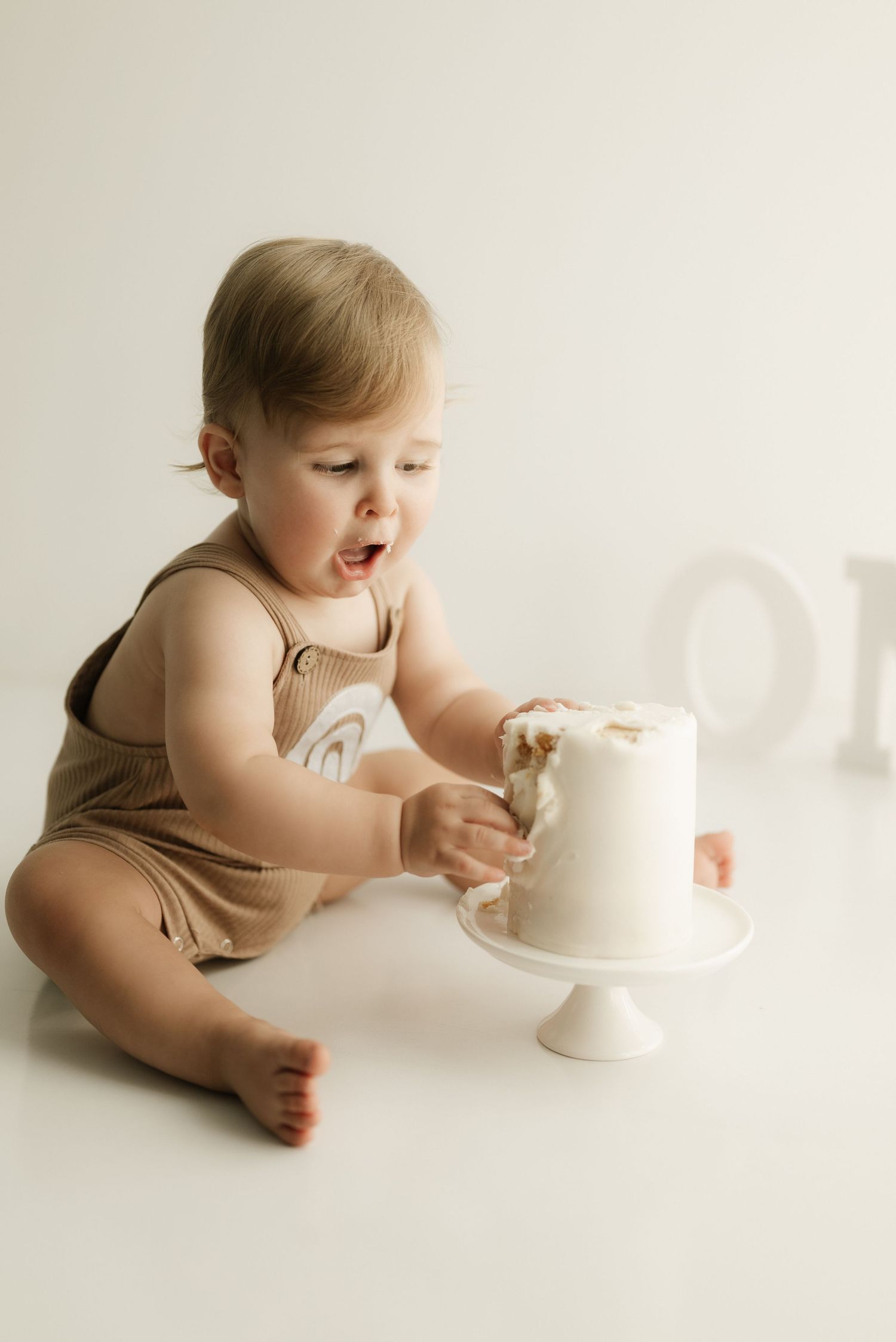 Baby in tan romper reaching for white birthday cake on pedestal.