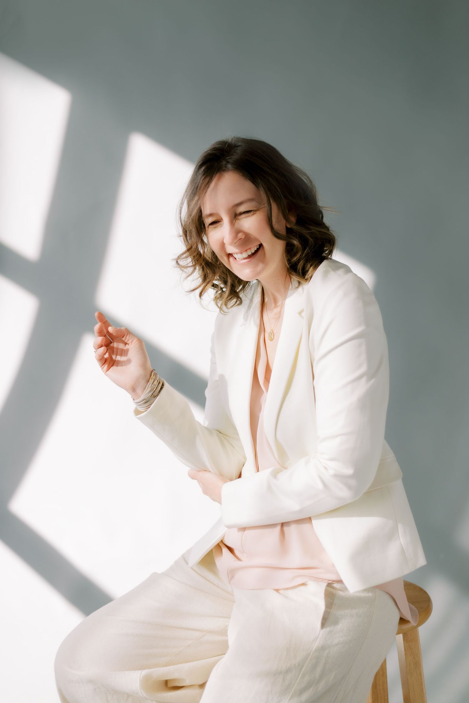 Person in white blazer laughs joyfully in studio with dramatic window lighting.