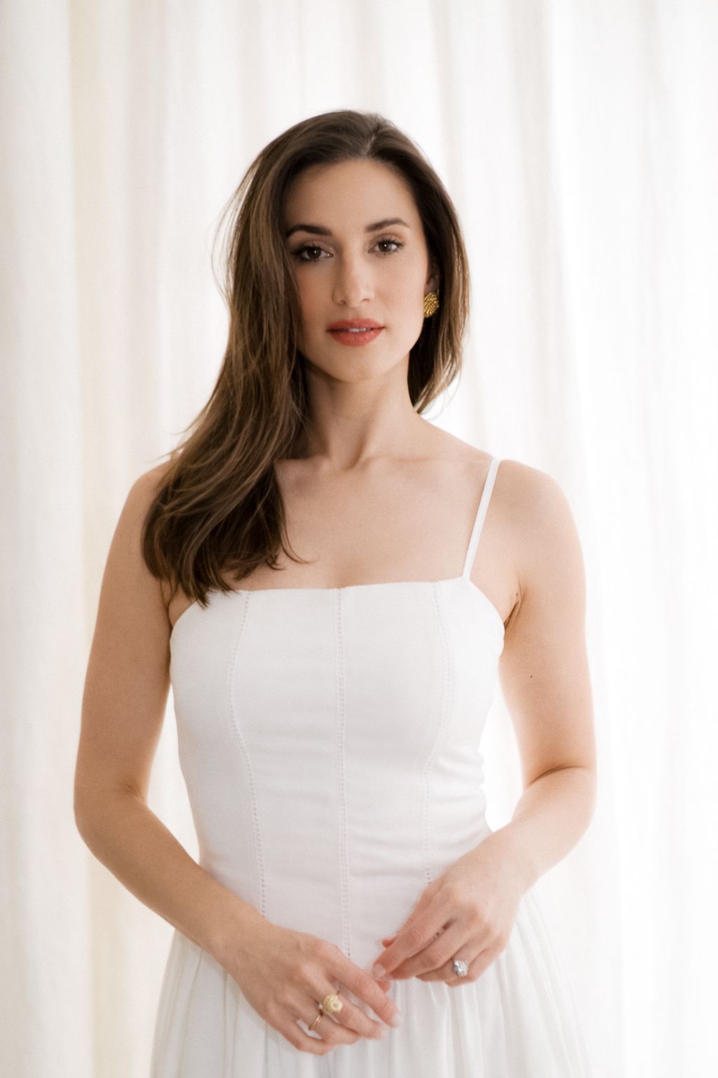 A person in a white spaghetti strap dress poses elegantly against a light curtain background in a soft, minimalist setting.