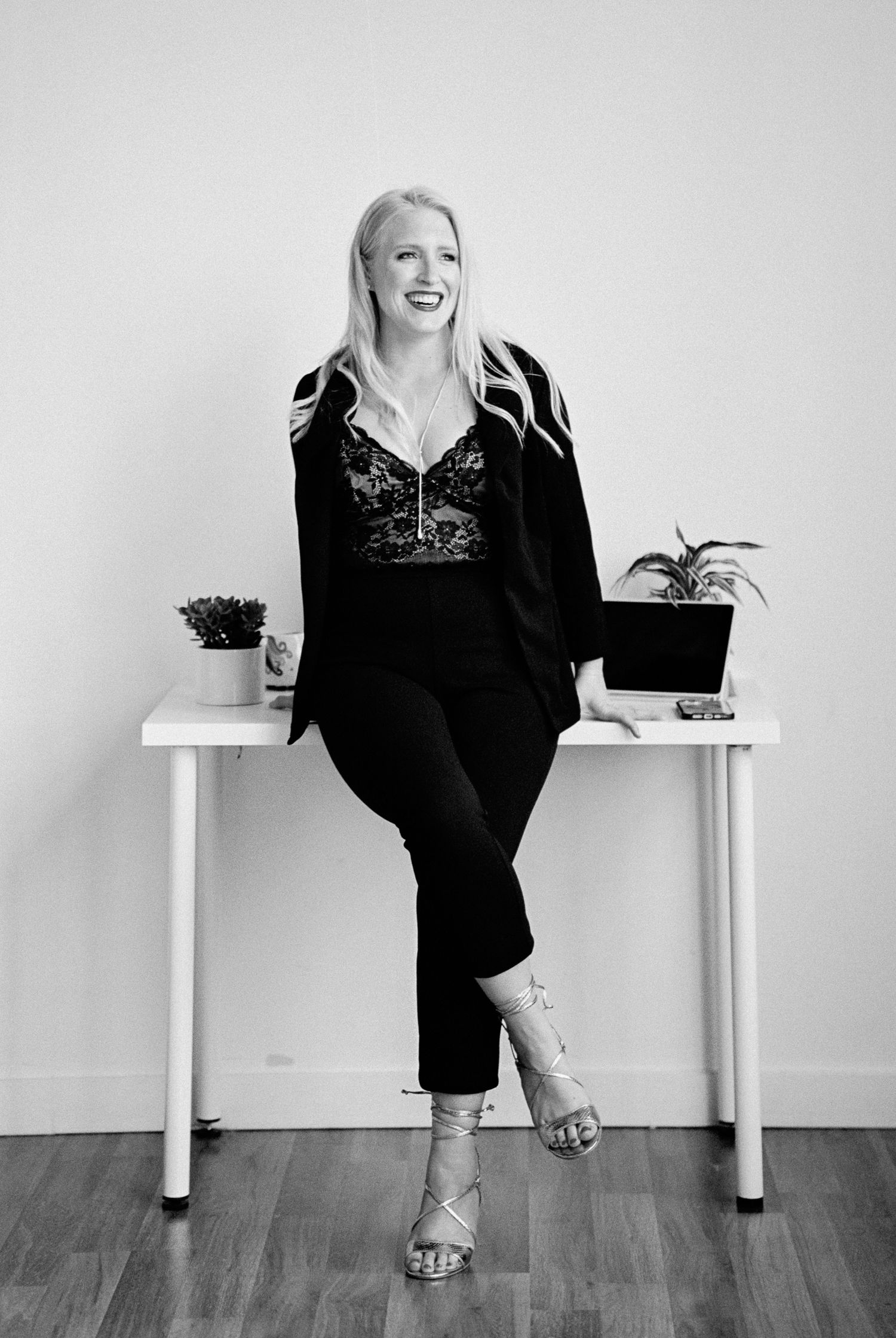 Professional woman in black outfit sits casually on white desk with plants.