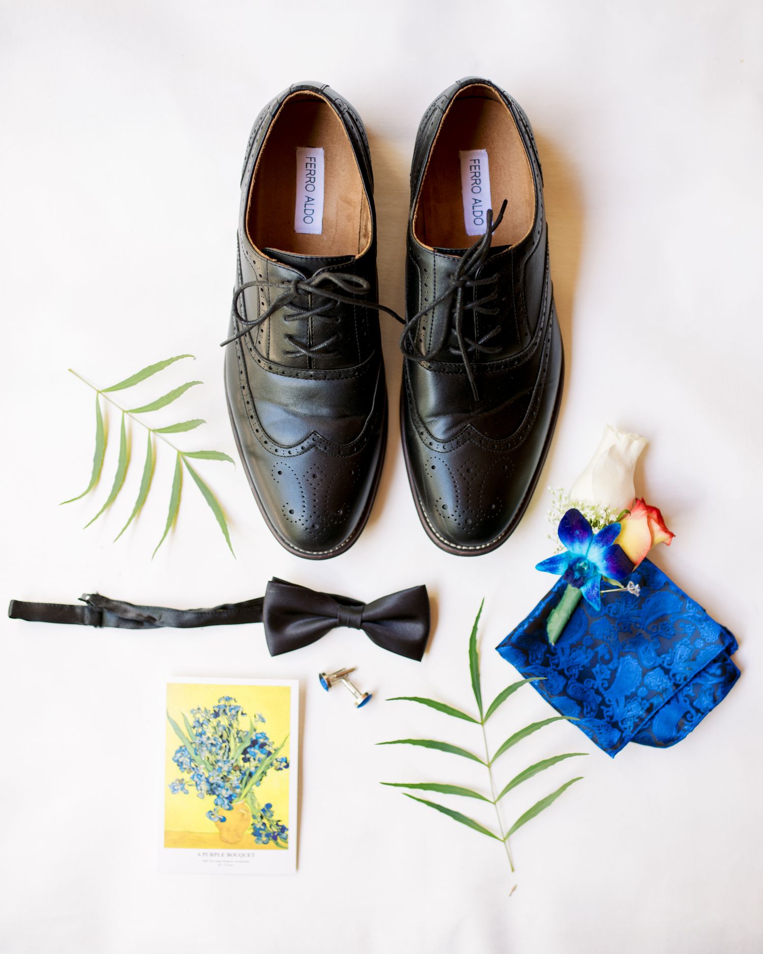Black wingtip dress shoes with wedding accessories including bowtie and blue pocket square on white background.