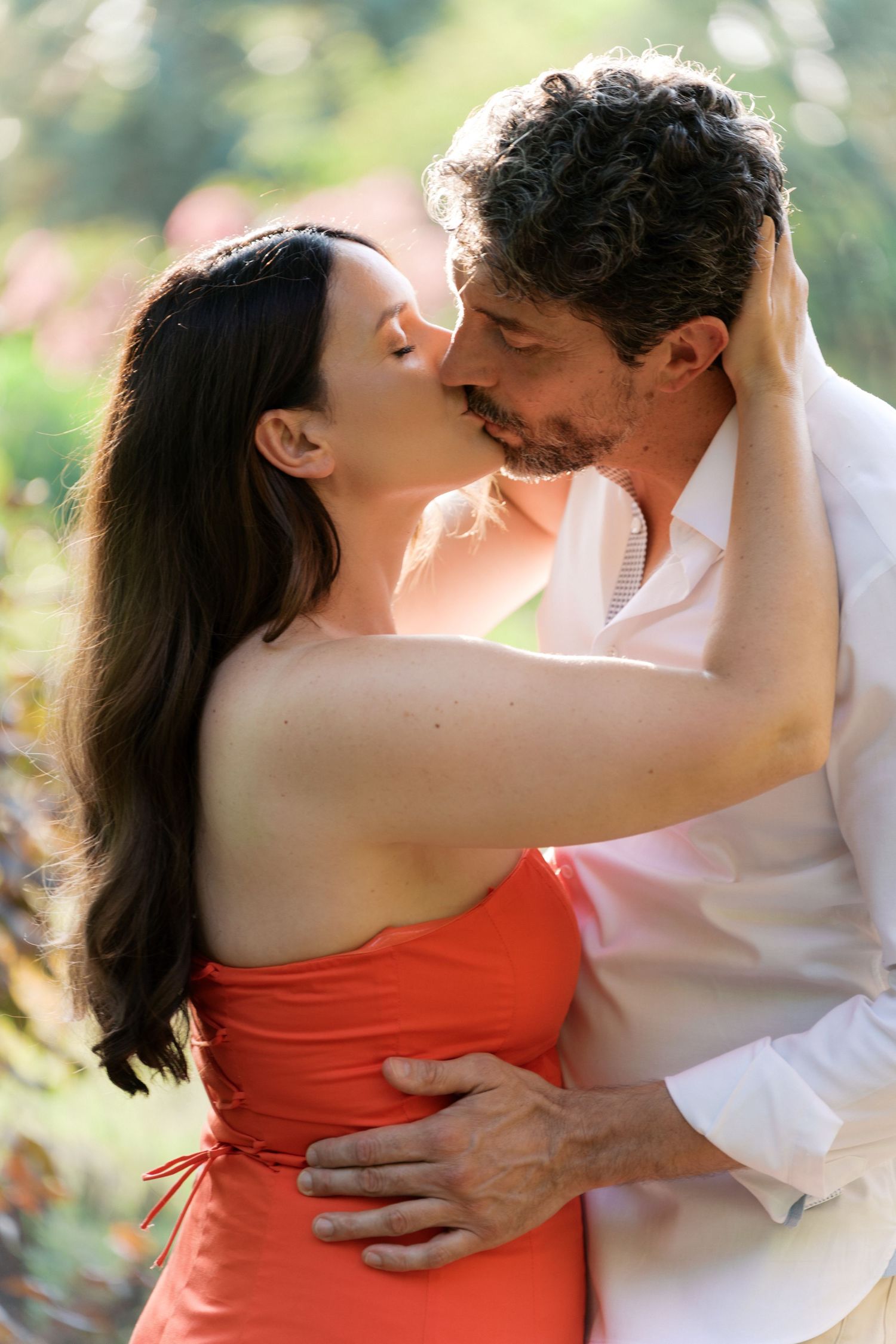 Romantic couple sharing a tender kiss outdoors, woman in red dress embracing man in white shirt amid lush greenery.