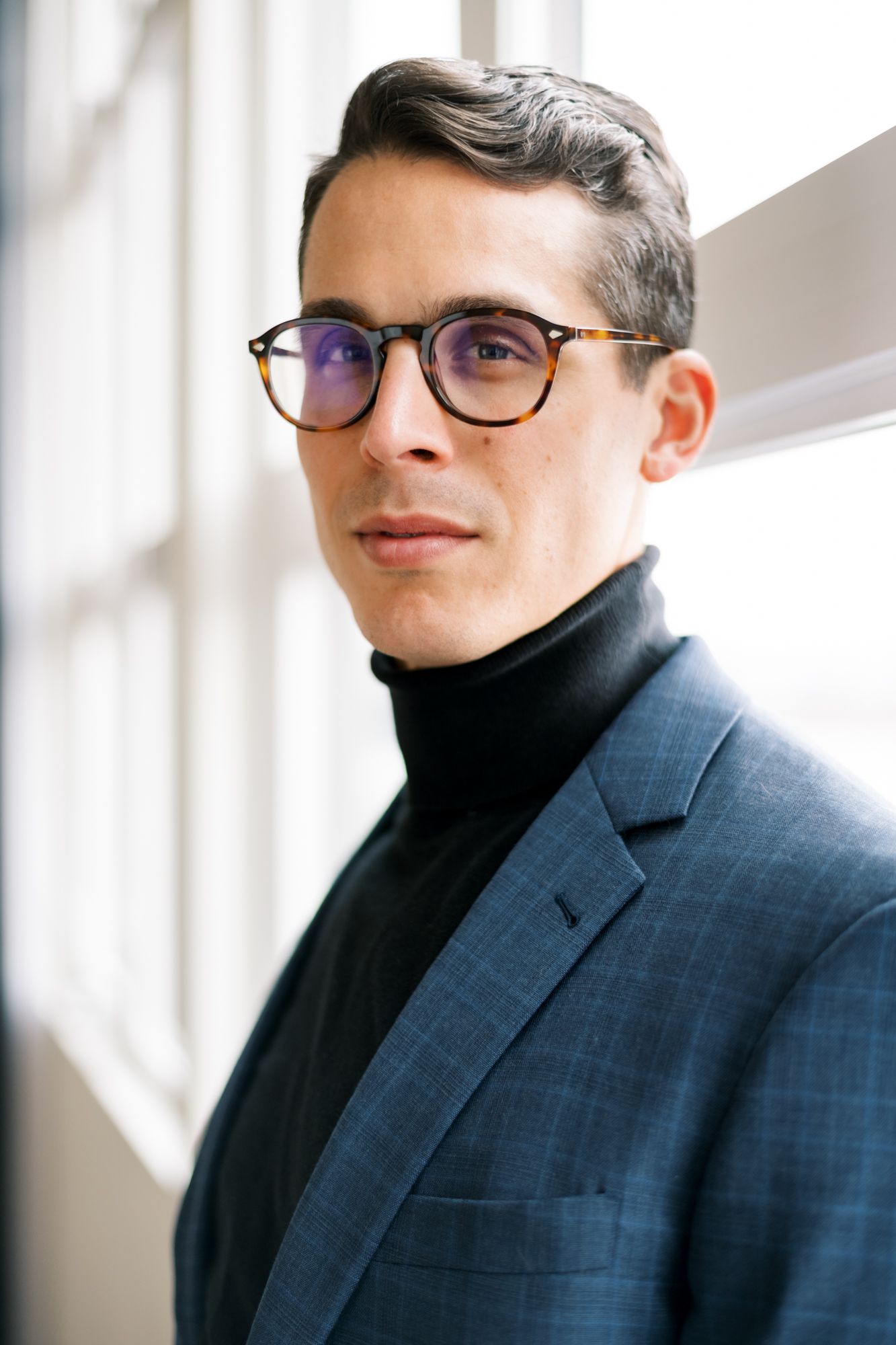 Professional headshot of person in turtleneck and glasses by window.