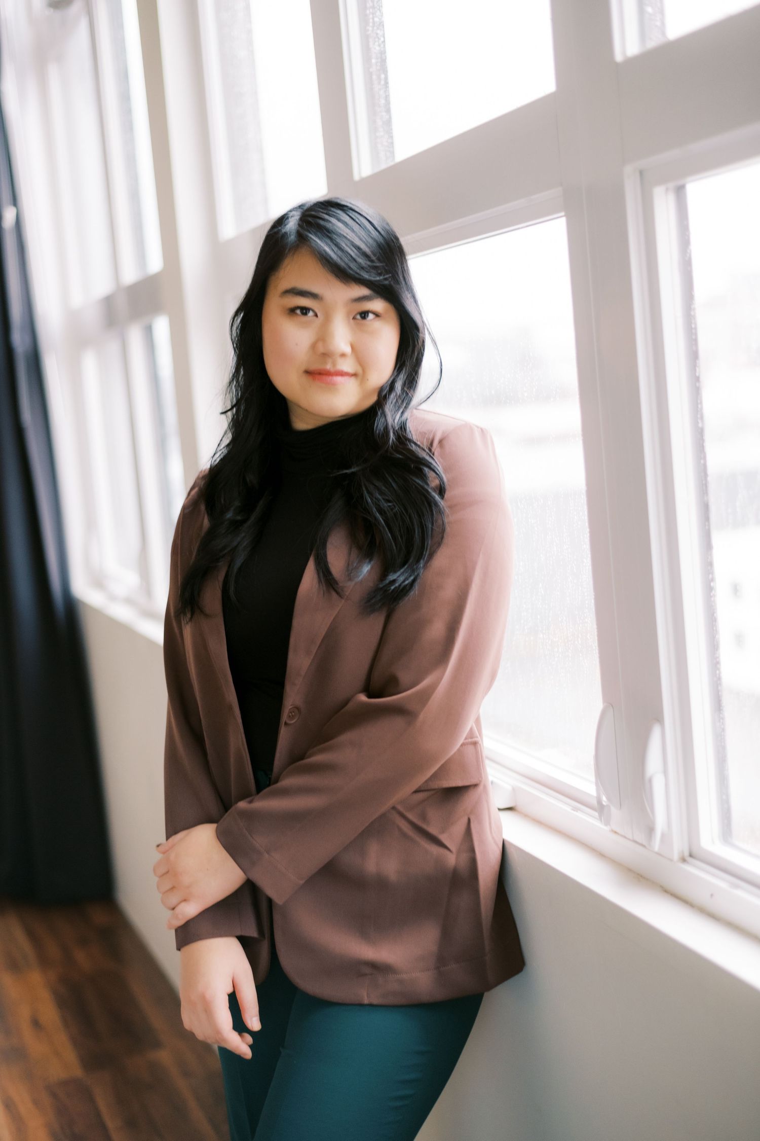 Professional portrait of person in brown blazer and turtleneck by window.