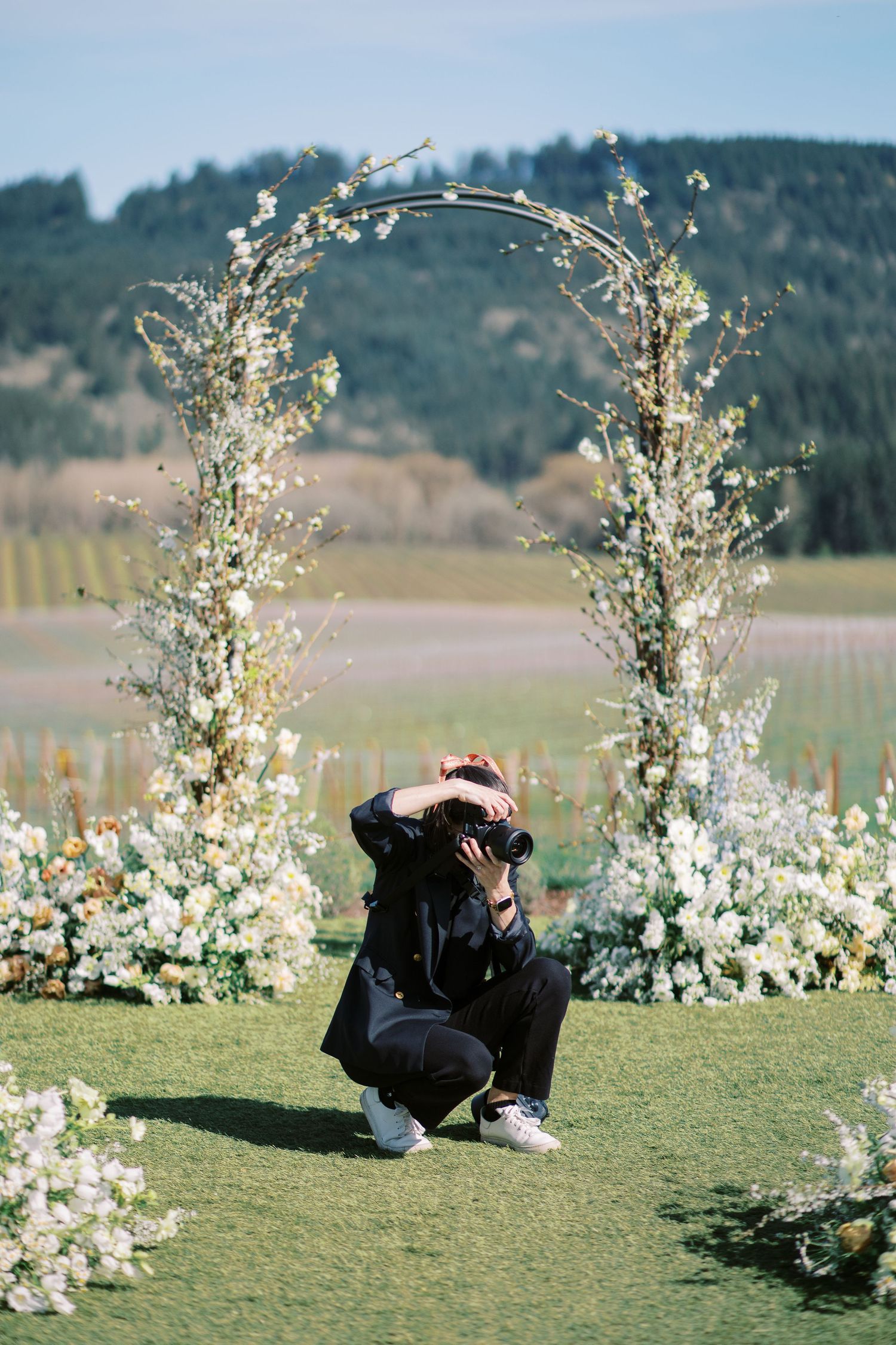 Photographer crouching under a floral arch with white blossoms against mountain backdrop.