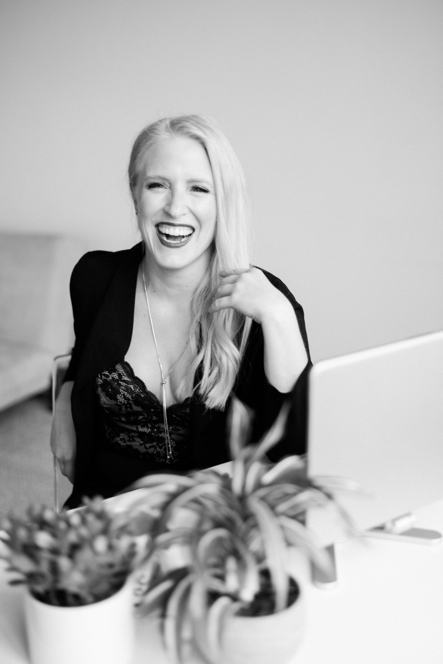 Black and white portrait of a joyful woman laughing while sitting near a potted plant.