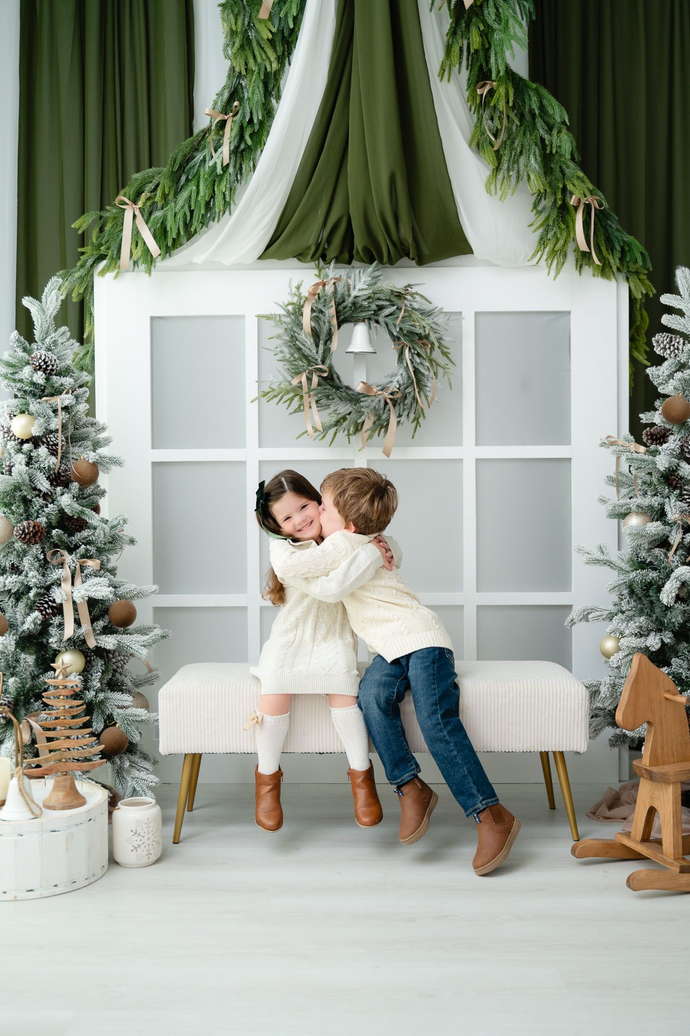 Two children hugging on a bench surrounded by snow-dusted Christmas trees in a festive white and green holiday setting.