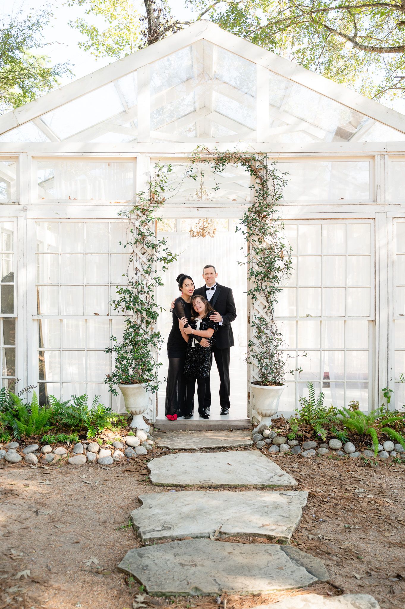 Family of three posing in formal attire under floral arch at elegant white greenhouse entrance with stone steps.
